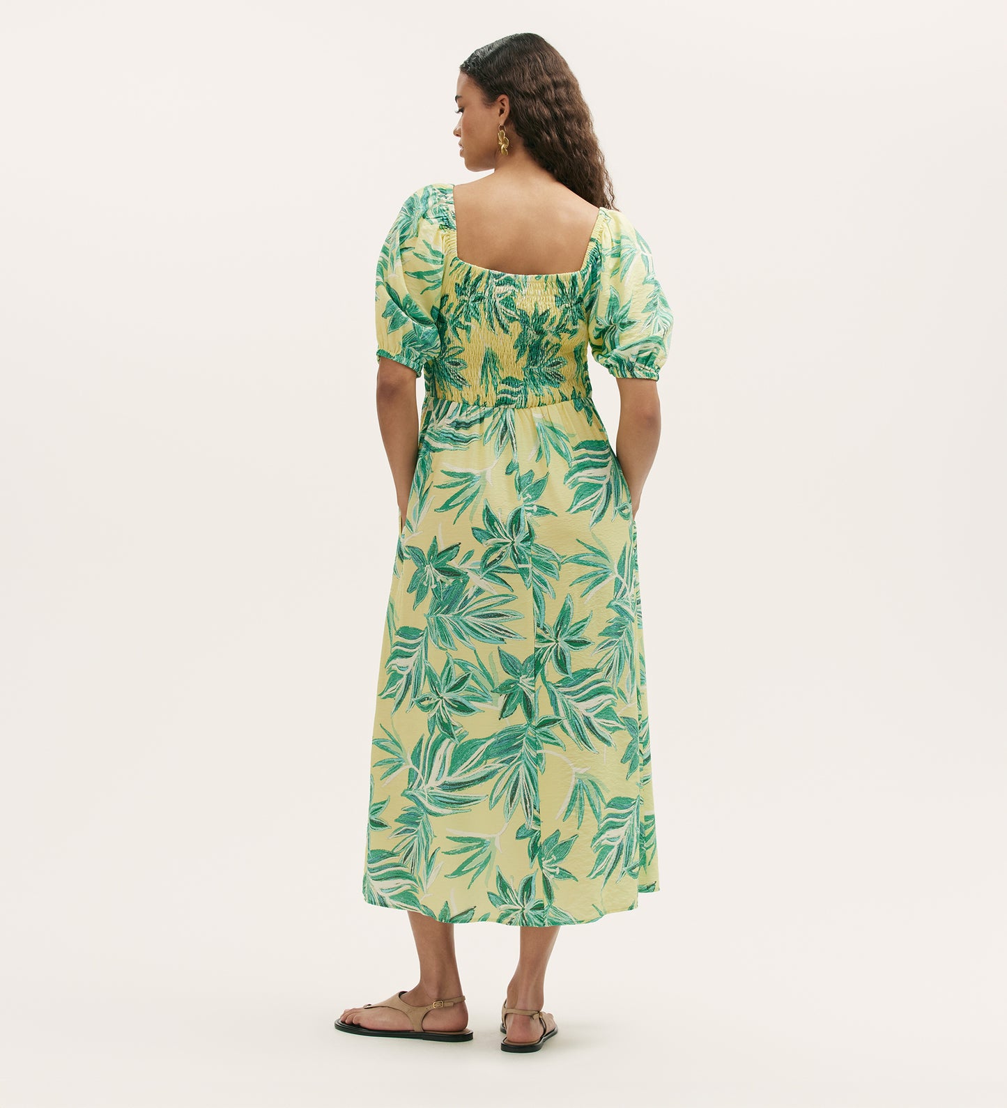 A woman stands facing away, wearing the Enzo Lime Tropical Bloom Midi Dress with green leaf prints, a square neckline, and short puff sleeves. Her wavy hair is down as she poses against a plain light background in sandals.