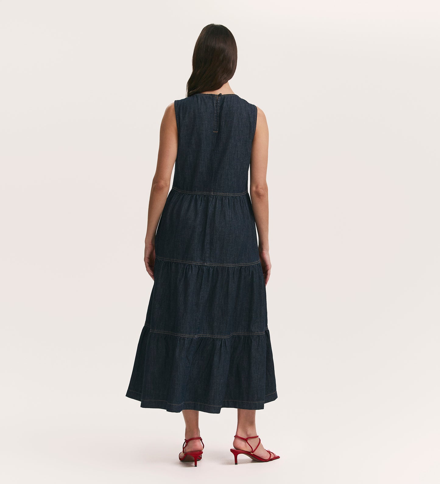 A woman with long brown hair, wearing the Essie Indigo Denim Sleeveless Midi Tiered Dress and red heeled sandals, stands facing away from the camera against a plain light background.