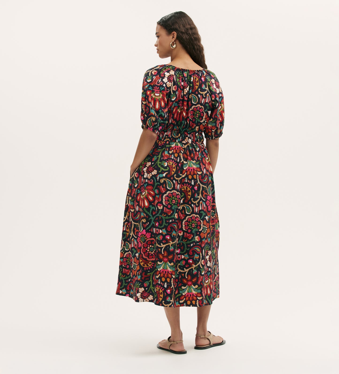 A woman with long braided hair and sandals faces away, wearing the Estee Black Floral Midi Smock Dress featuring a colorful floral pattern, fit-and-flare silhouette, midi length, and short puff sleeves against a plain light background.