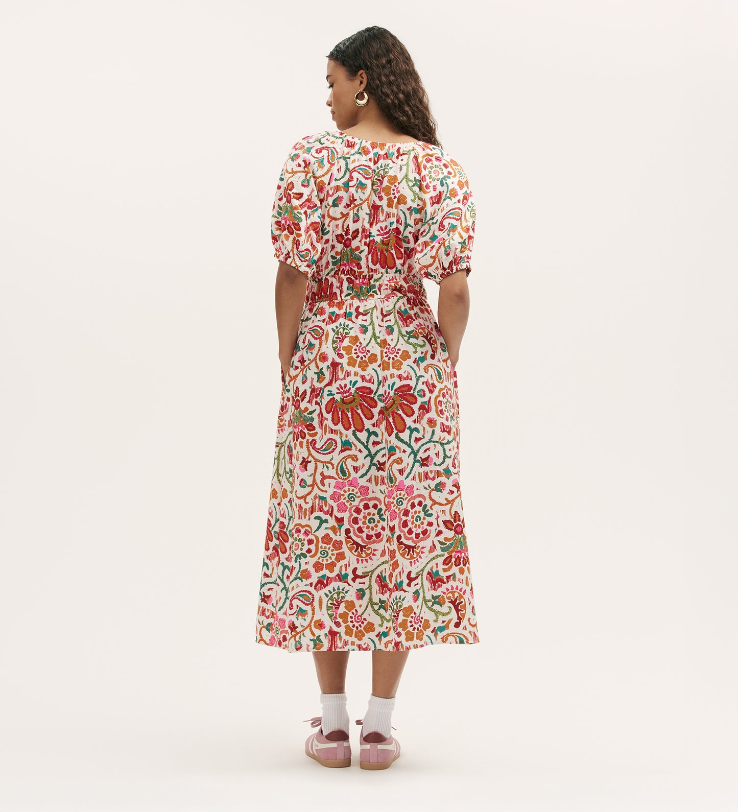 A woman with long curly hair wears the Estee Ivory Floral Midi Smock Dress, featuring puffed sleeves and a colorful floral pattern, paired with pink sandals and earrings. She stands against a plain white background.