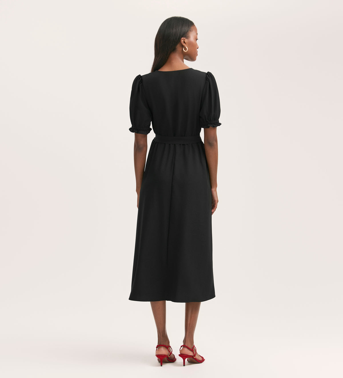 A woman faces away, wearing the Everleigh Black Crepe Midi Dress with short puff sleeves, a v-neck, and a belt, paired with red heels against a plain light background.