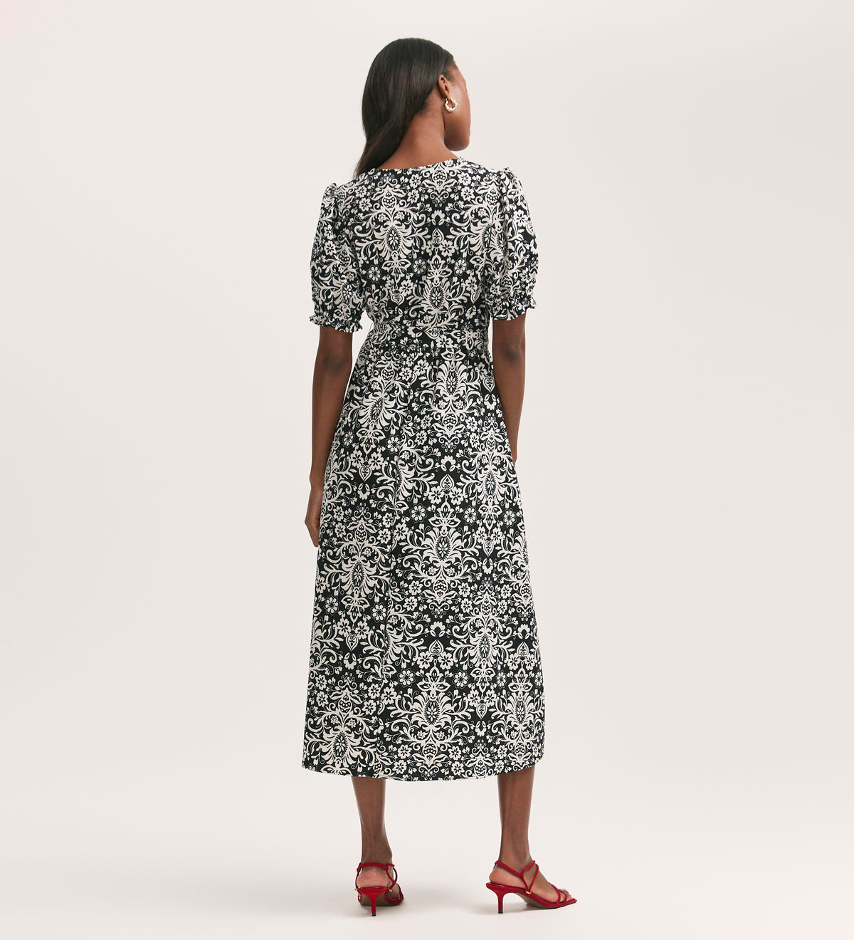 A woman faces away in the Everleigh Black Paisley Crepe Midi Dress featuring puffed sleeves. Her long dark hair contrasts with her red heeled sandals against a plain, light-colored background.