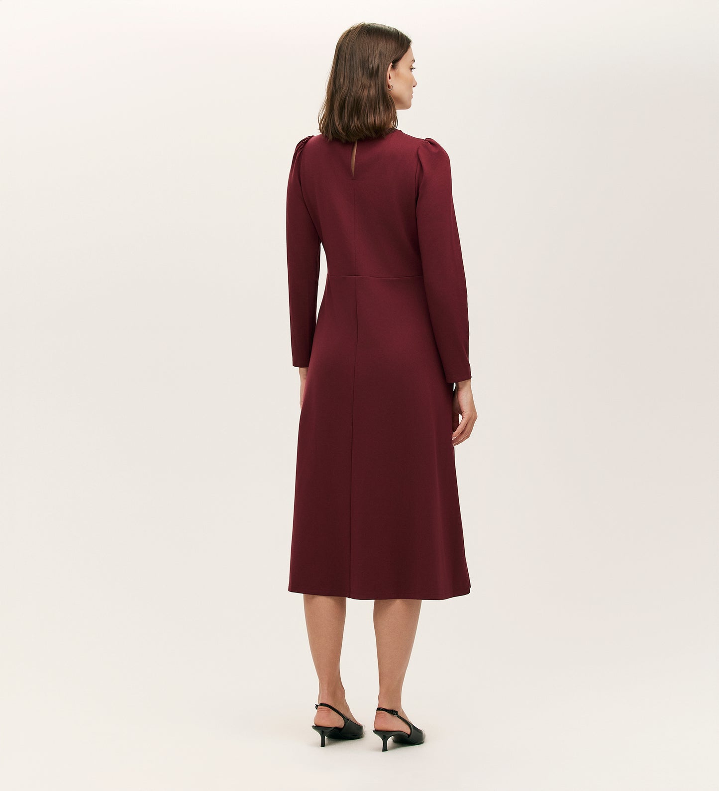 A woman stands facing away, wearing the Felice Burgundy Ponte Jersey Midi Dress with a tailored silhouette and black heeled sandals against a plain light background.