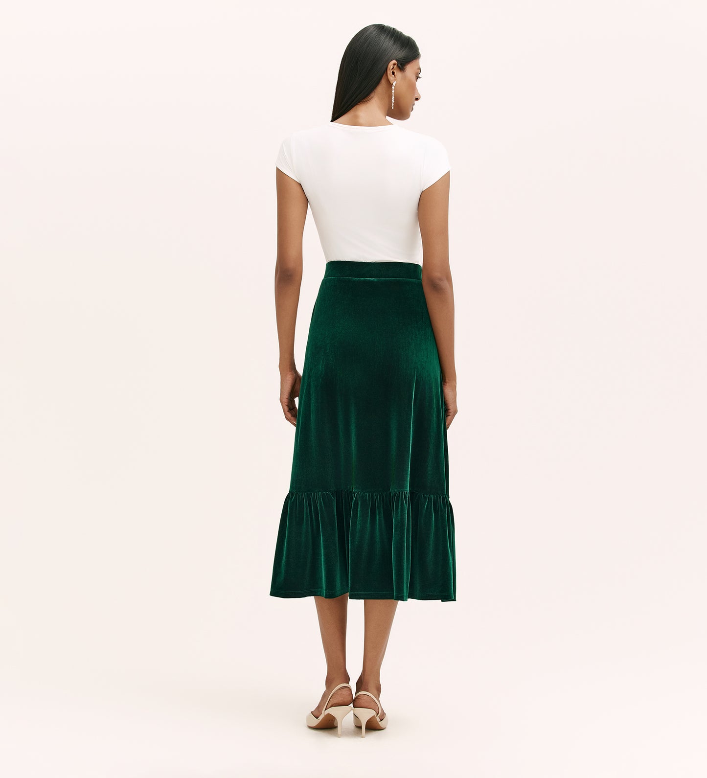 A woman with long, straight hair poses in a neutral studio, wearing the Joline Bottle Green Velvet Midi Skirt with an elasticated waistband, paired with a white short-sleeve top and beige heeled sandals.