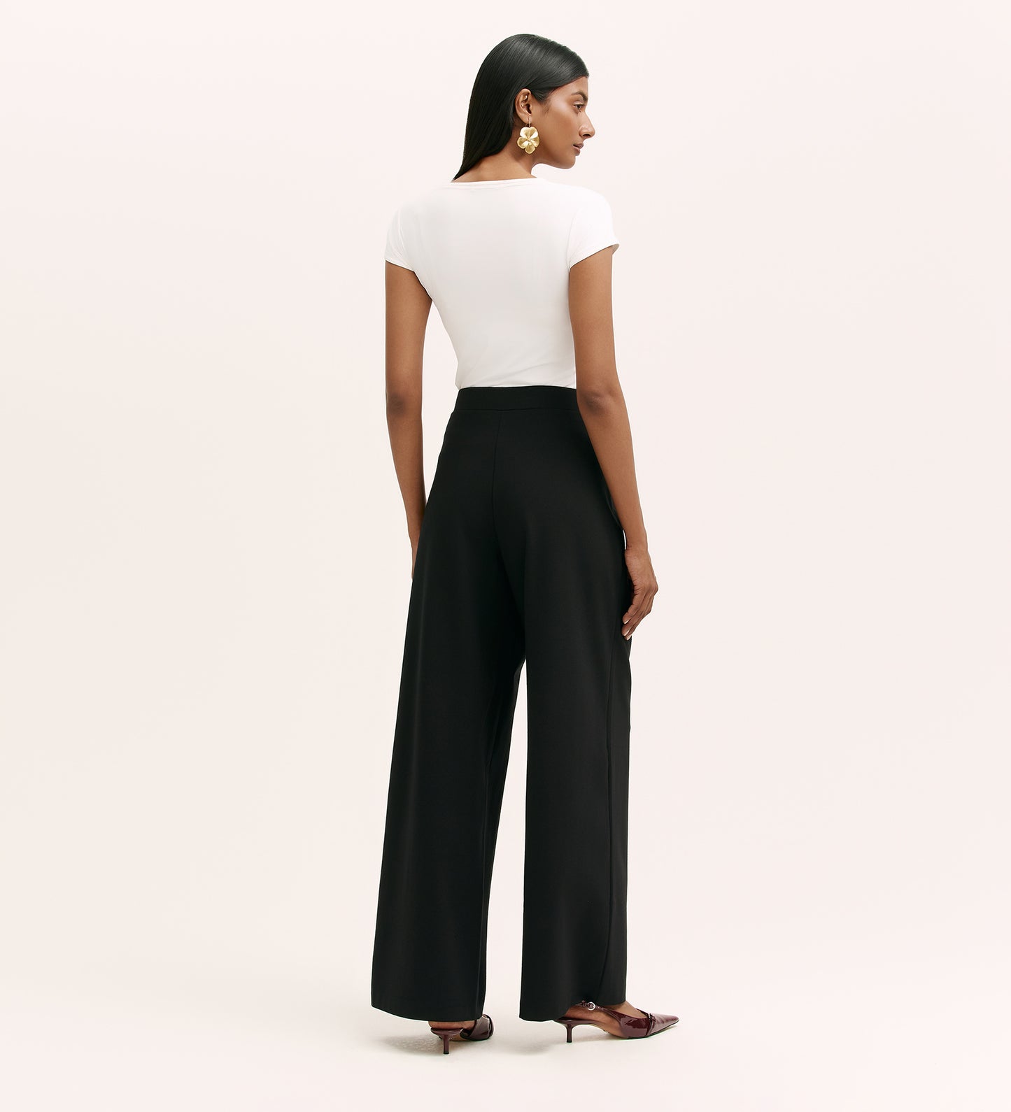 A woman faces away, wearing a fitted white short-sleeve top, the Kenzie Black Ponte Jersey Wide Leg Trousers with an elastic waistband, pointed-toe brown shoes, and large gold statement earrings against a plain background.