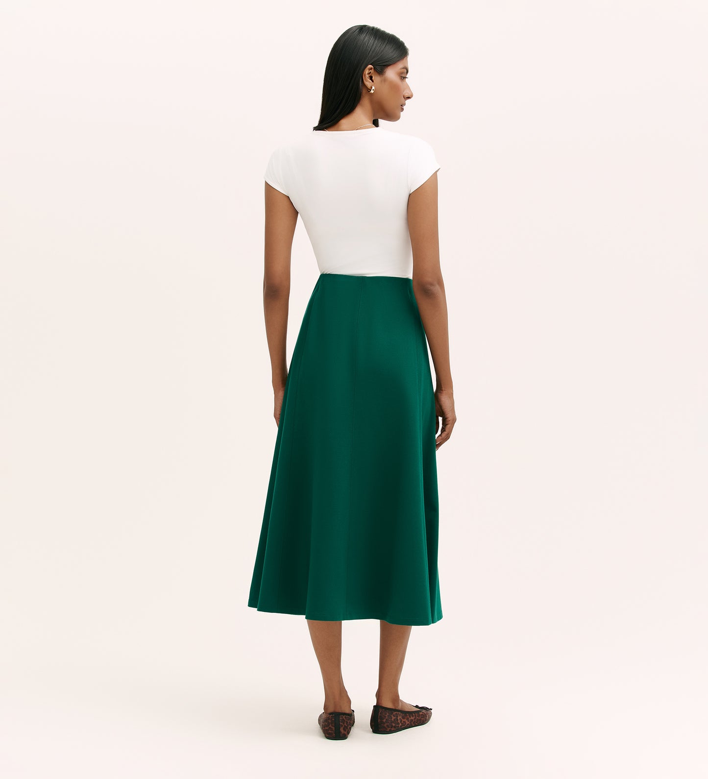 A woman with straight dark hair faces away, wearing a short-sleeved white top, black flats, and the Mae Bottle Green Ponte Jersey Midi Skirt with an elasticated waistband, set against a plain light background.