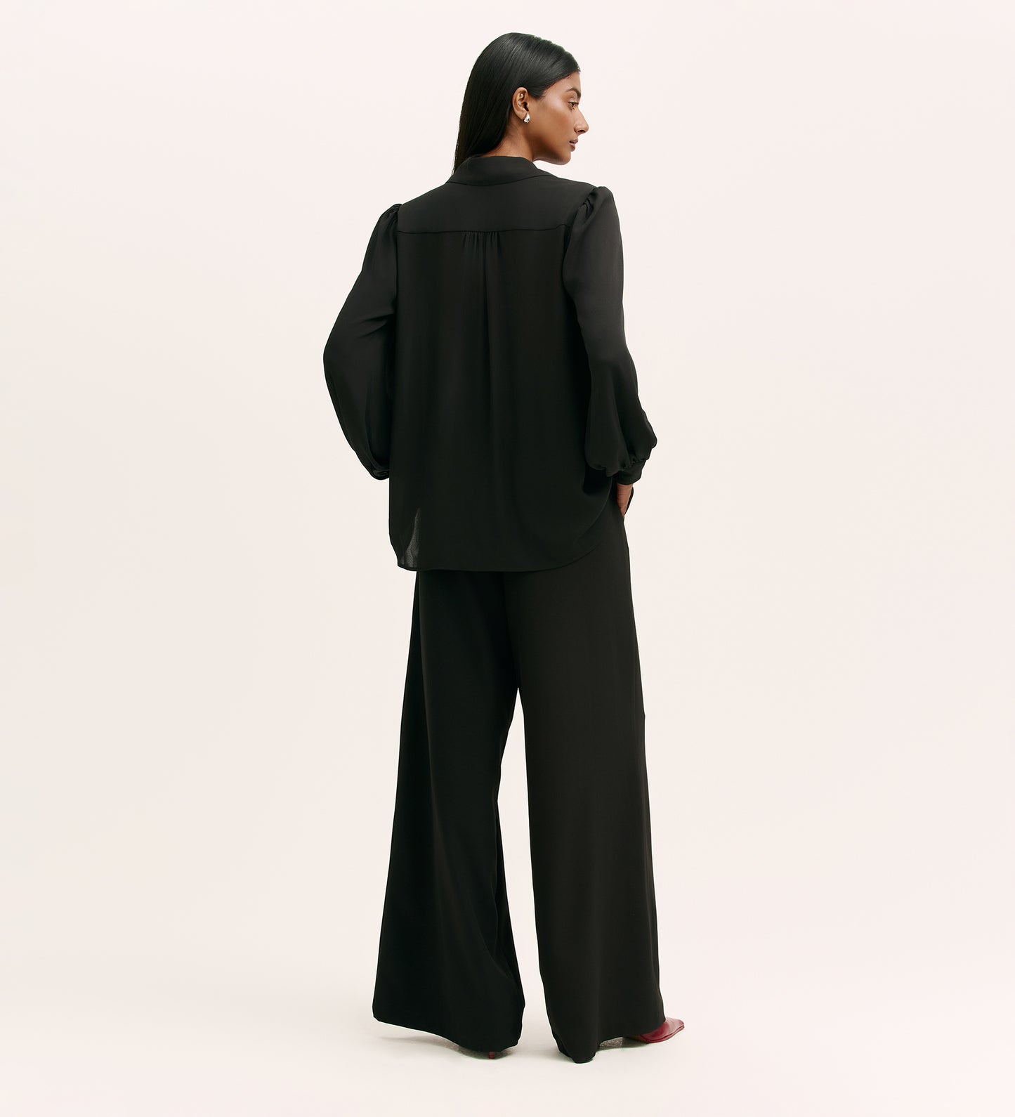 A person with long dark hair stands facing away, wearing the Melina Black Crepe Blouse and wide-leg black trousers against a plain light background.