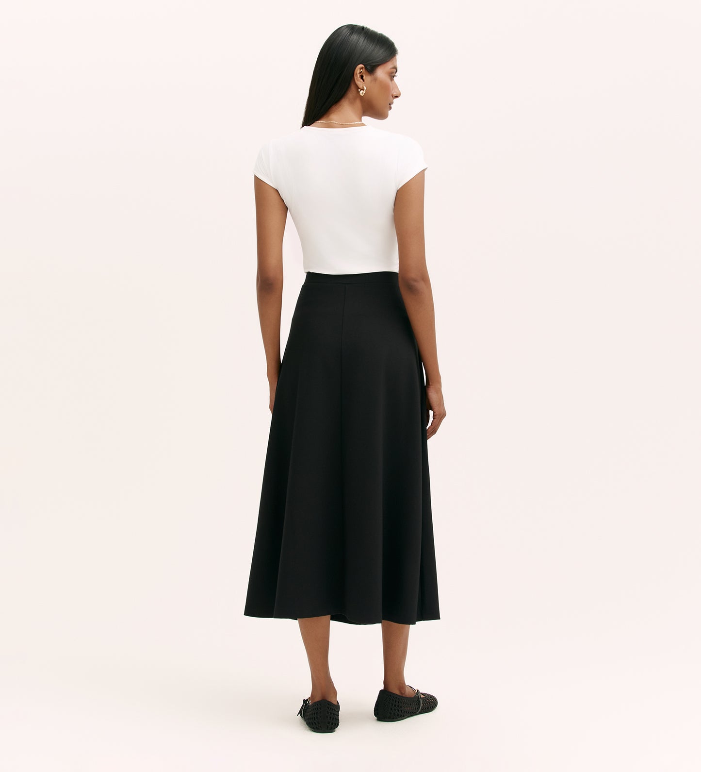 A woman with straight dark hair faces away, wearing a short-sleeved white top and the Ruth Black Ponte Jersey Midi Skirt, which is high-waisted with an elastic waistband. She pairs it with black flats against a plain light background.