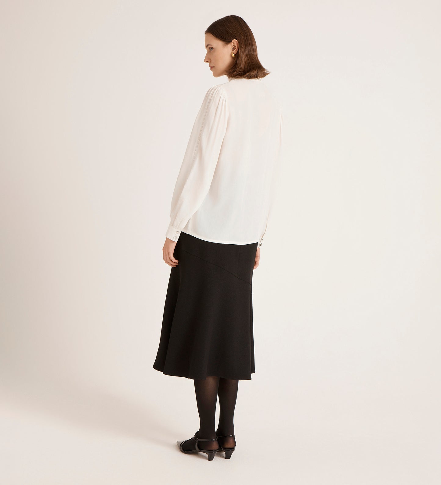 A woman, back turned, wears the Flora Ivory Crepe Blouse with a black midi skirt, tights, and shoes against a plain light background.