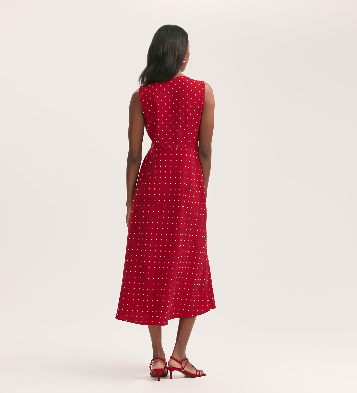 A woman faces away, wearing the Flora Red Polka Dot Midi Dress in crease-free crepe fabric with red heeled sandals, set against a plain light background.