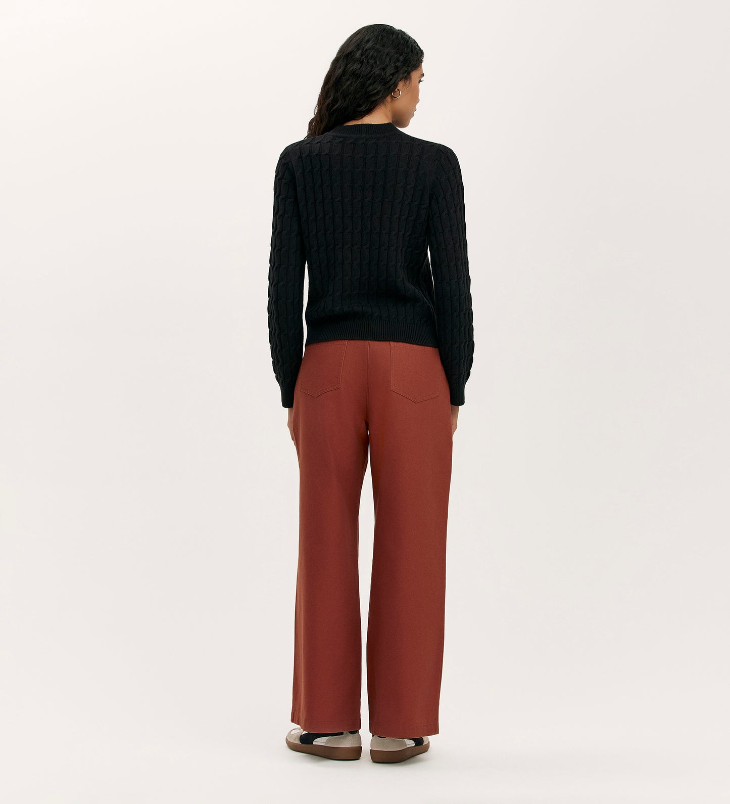 A woman with long, wavy dark hair faces away, wearing the Frida Black Knitted Jumper and loose rust-colored pants. Brown shoes complement her outfit, while a light background highlights this cosy wardrobe essential.