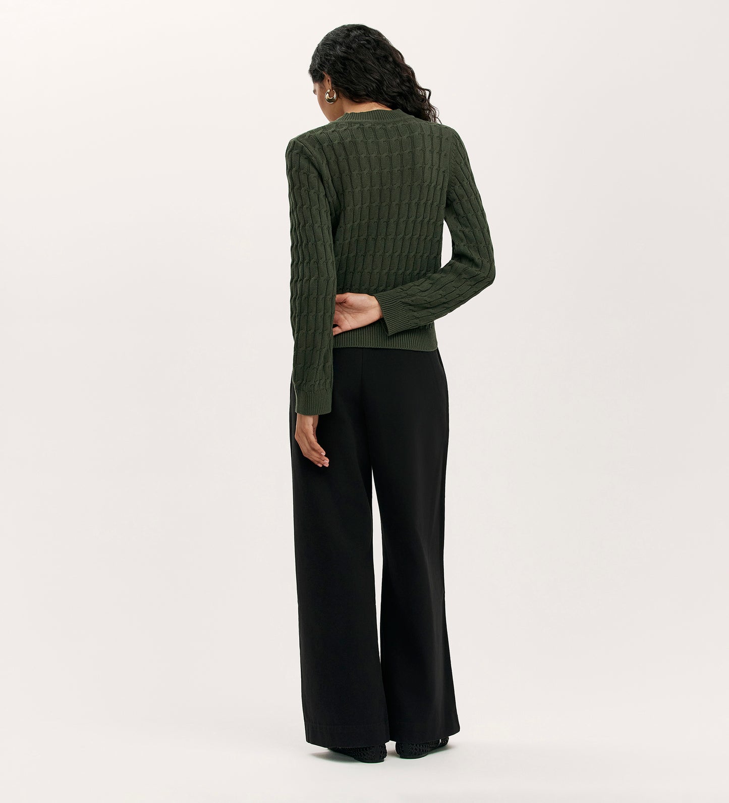 A person with long wavy hair, wearing the Frida Forest Green Knitted Jumper and loose black pants, stands facing away with one hand behind their back against a plain light background.
