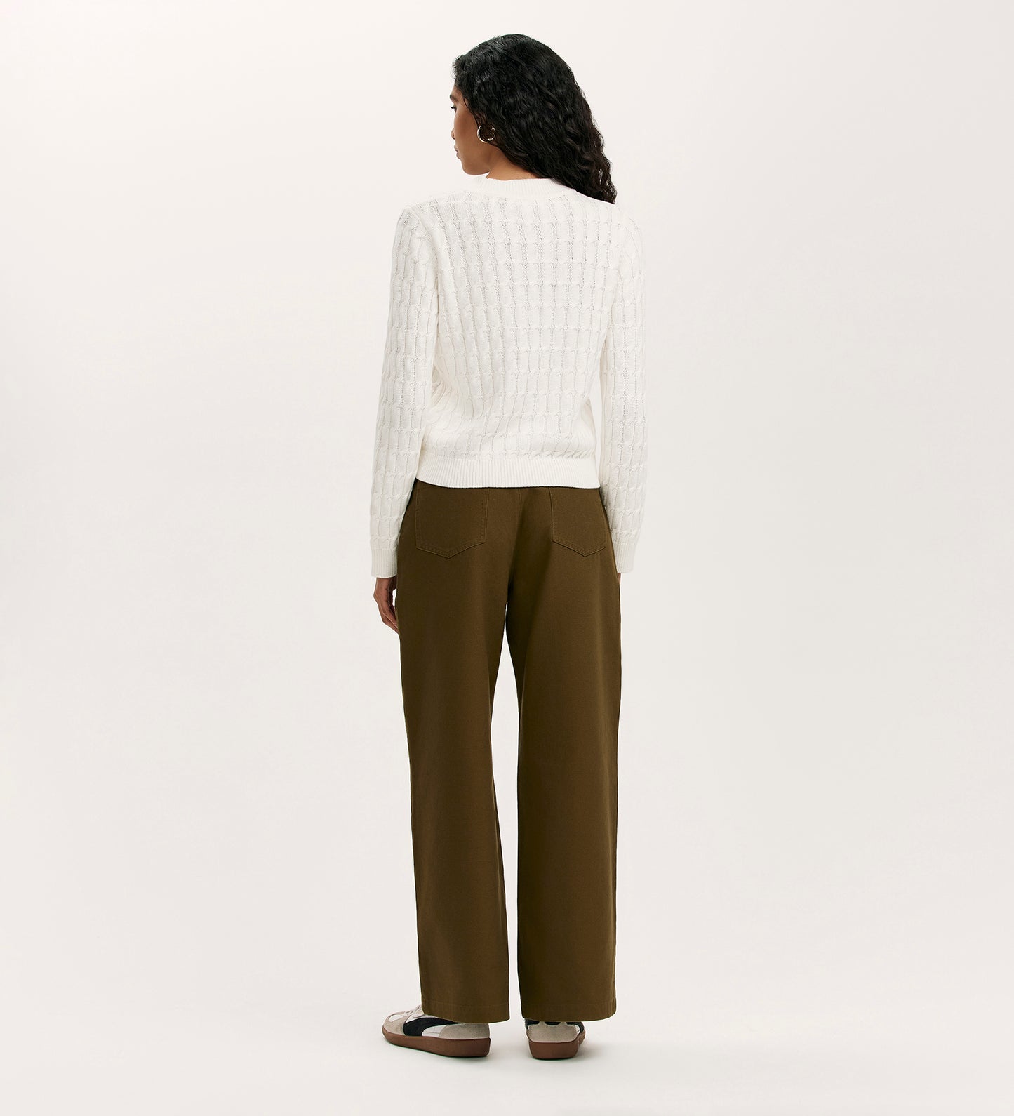 A person with long dark hair stands facing away, wearing the Frida Ivory Knitted Jumper, olive green wide-leg pants, and brown sandals against a plain light background.