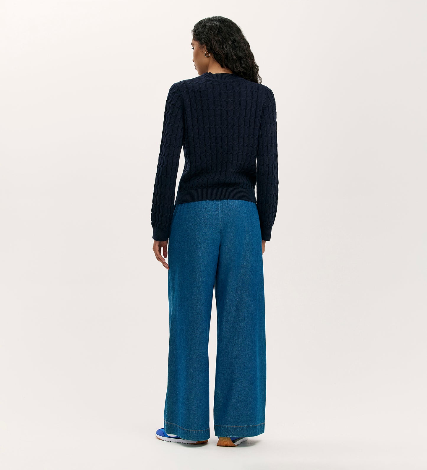 A person with long curly hair faces away, wearing the cosy Frida Navy Knitted Jumper paired with wide-leg blue jeans and sneakers against a plain light background.