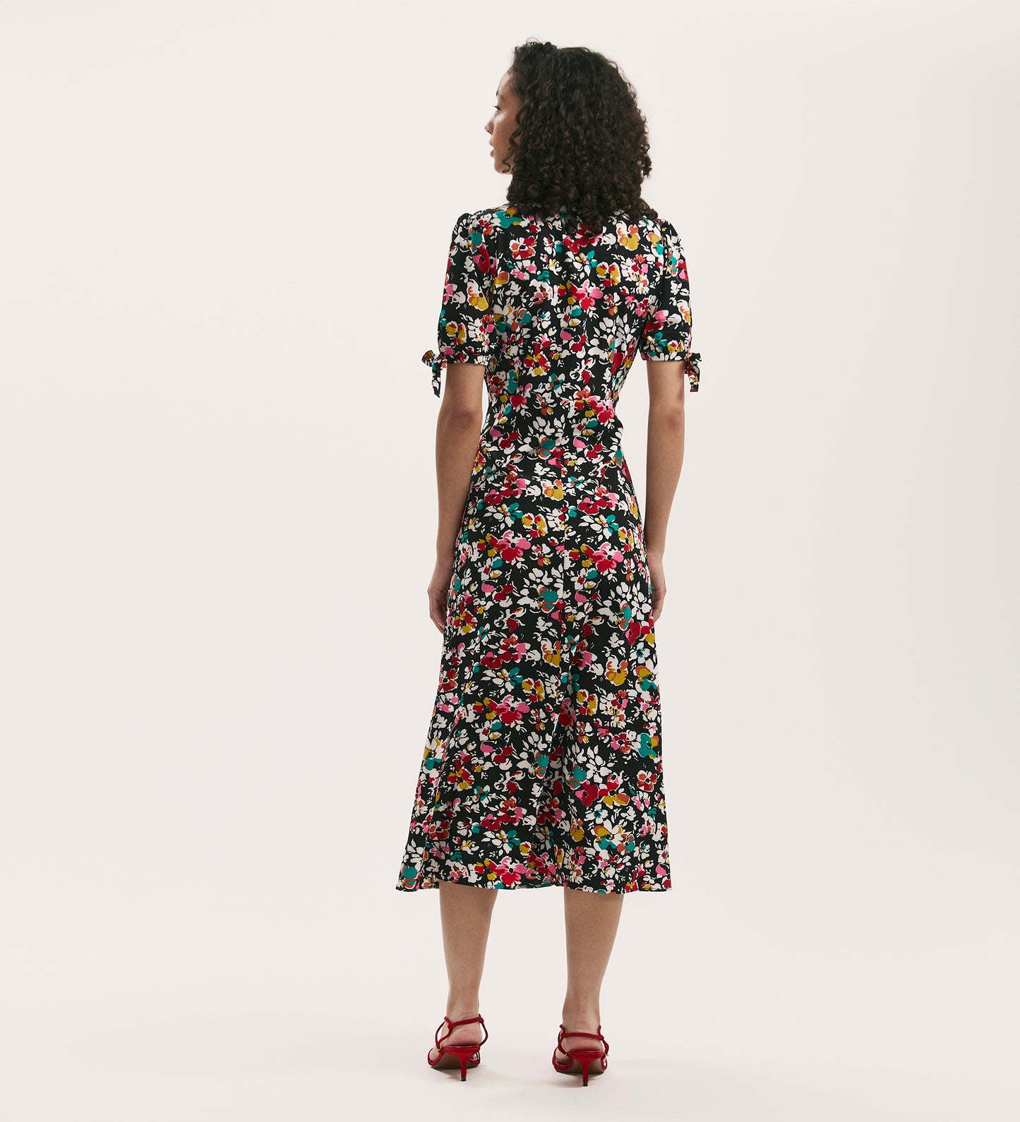 Seen from behind against a white background, a woman with curly hair wears the Gabriella Black Floral Crepe Midi Tea Dress with short puff sleeves and red heeled sandals, crafted from crease-free crepe fabric.