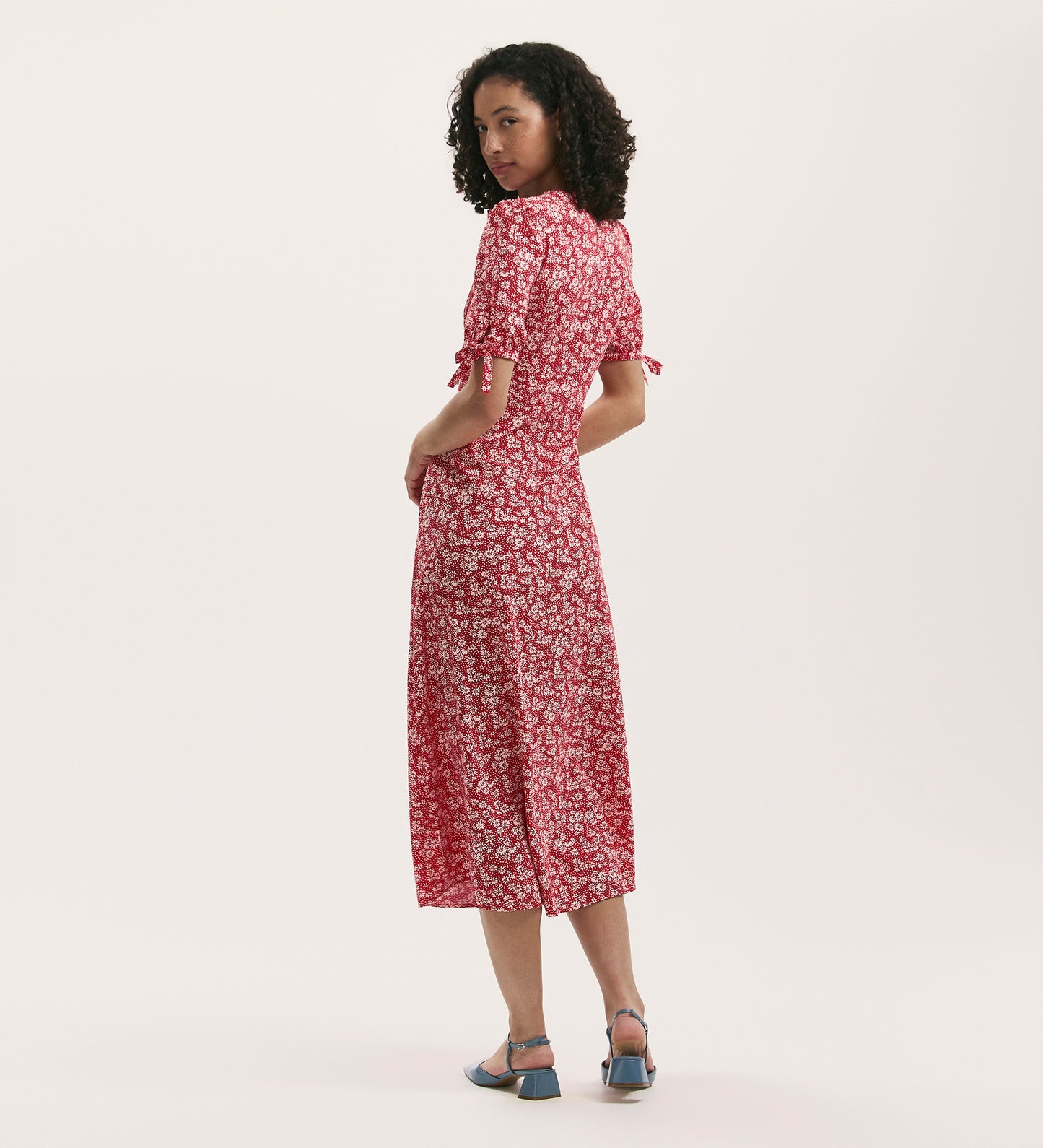 A woman with curly hair models the Gabriella Red Daisy Crepe Midi Tea Dress, a crease-free, V-neck style with short sleeves. She stands partially turned from the camera, glancing over her shoulder in light blue low-heeled sandals.