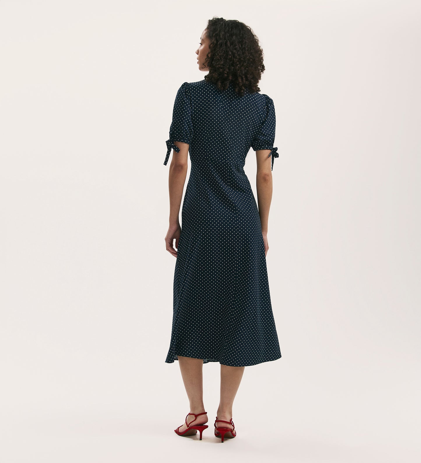 A woman with curly hair faces away, wearing the Gabriella Navy Polka Dot Crepe Midi Tea Dress and red heeled sandals against a plain light background.