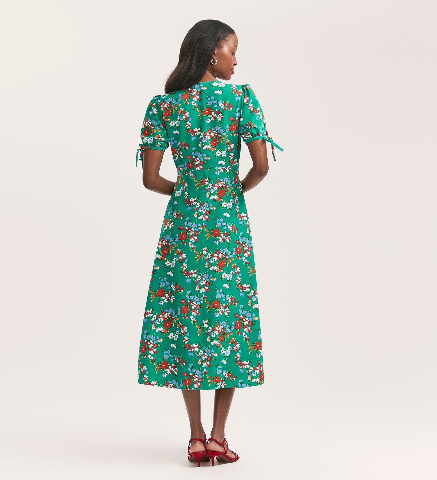 A woman, seen from behind against a light gray background, wears the Gabriella Green Floral Crepe Midi Dress with a V-neck, fitted waist, short sleeves, and pairs it with red strappy heels.