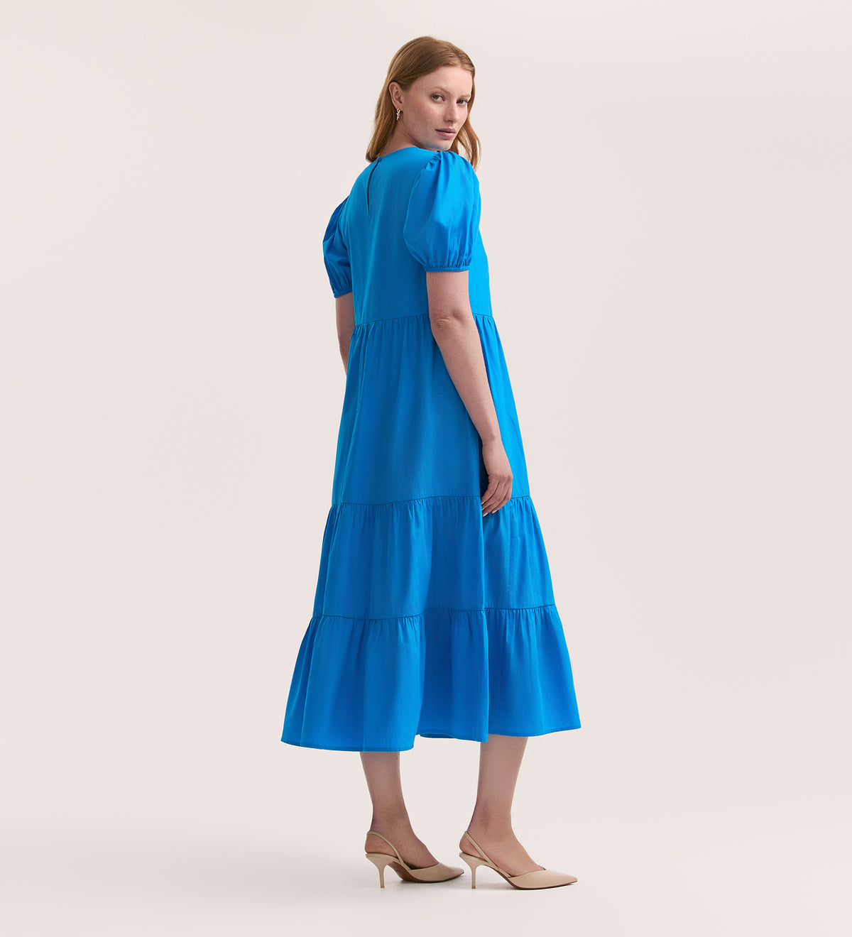 A woman stands turned to the side, looking over her shoulder, wearing the Genevive Blue Cotton Midi Dress with a tiered skirt and nude heels. Her straight light brown hair contrasts against a plain, light background.