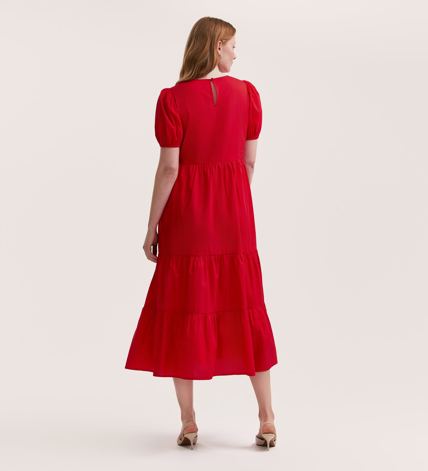 A woman with light brown hair faces away, wearing the Genevive Red Cotton Midi Dress with short sleeves and beige heeled sandals, set against a plain light background.