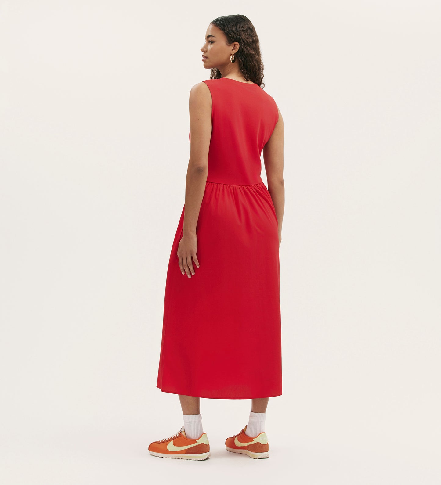 A woman faces away in a Georgie Red Cotton Sleeveless Midi Dress, a relaxed fit that falls below the knees, paired with orange Nike sneakers and white socks against a plain light background.