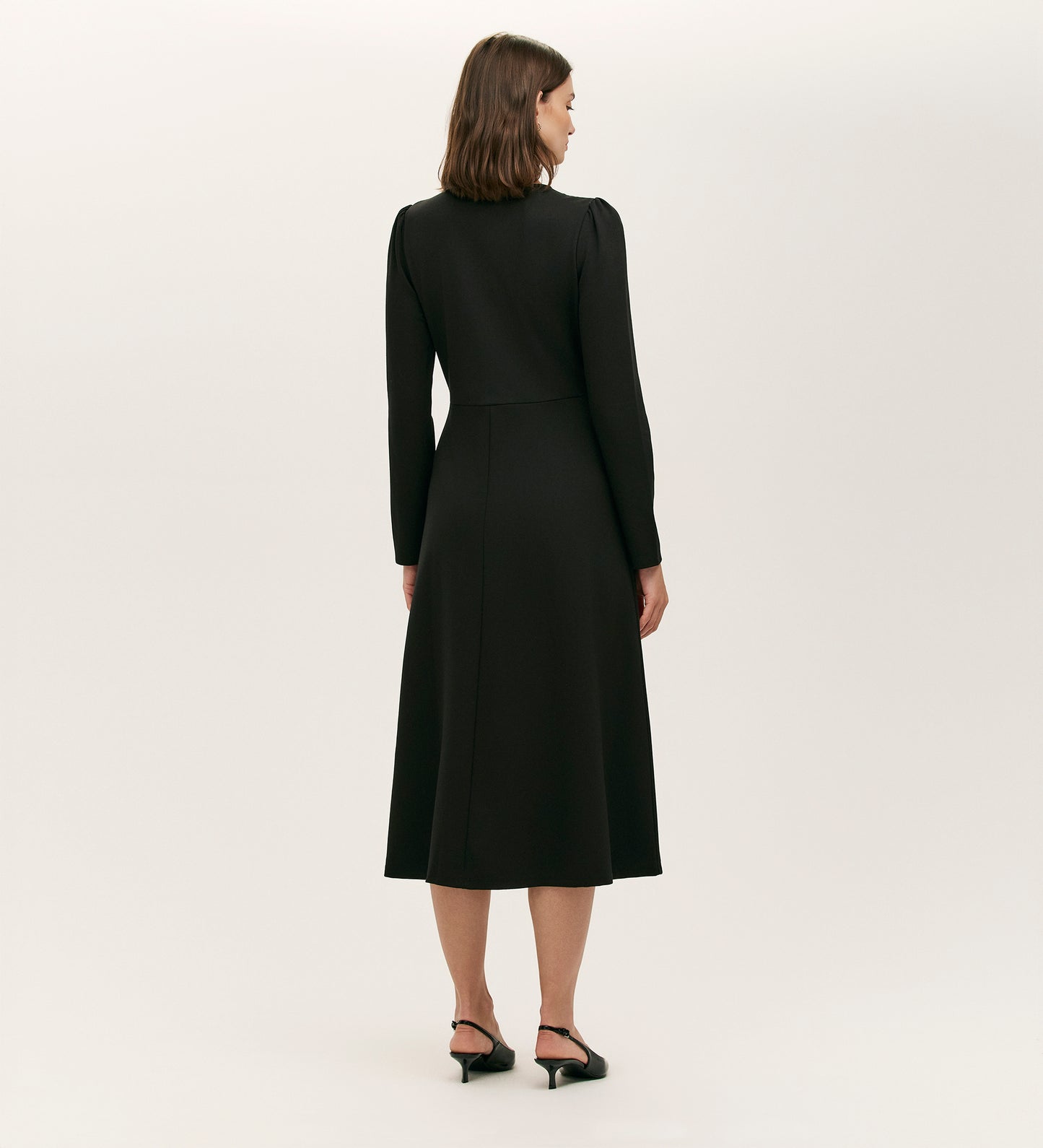 A woman with shoulder-length brown hair stands facing away, wearing the Gilly Black Ponte Jersey Midi Dress and black low-heeled shoes against a plain light background.