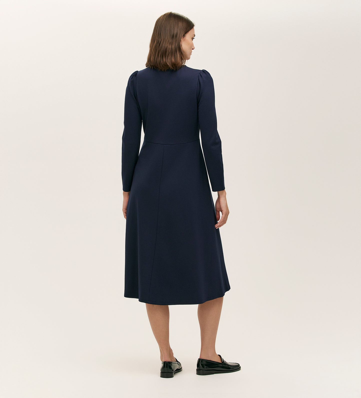 A woman faces away from the camera wearing the Gilly Navy Ponte Jersey Midi Dress with long sleeves and black loafers, set against a plain light background.