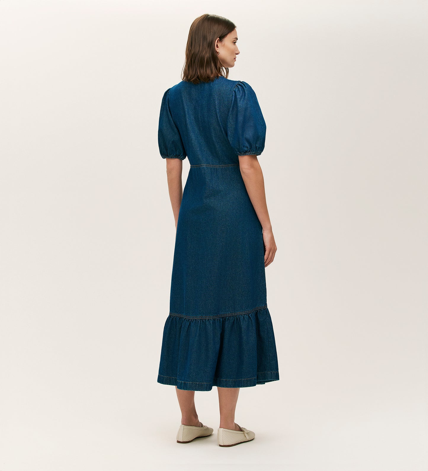 A woman with shoulder-length brown hair and beige slip-on shoes faces away, wearing the Goldie Indigo Denim Midi Dress featuring puffed sleeves, a ruffled hem, and button-through front, set against a plain light background.