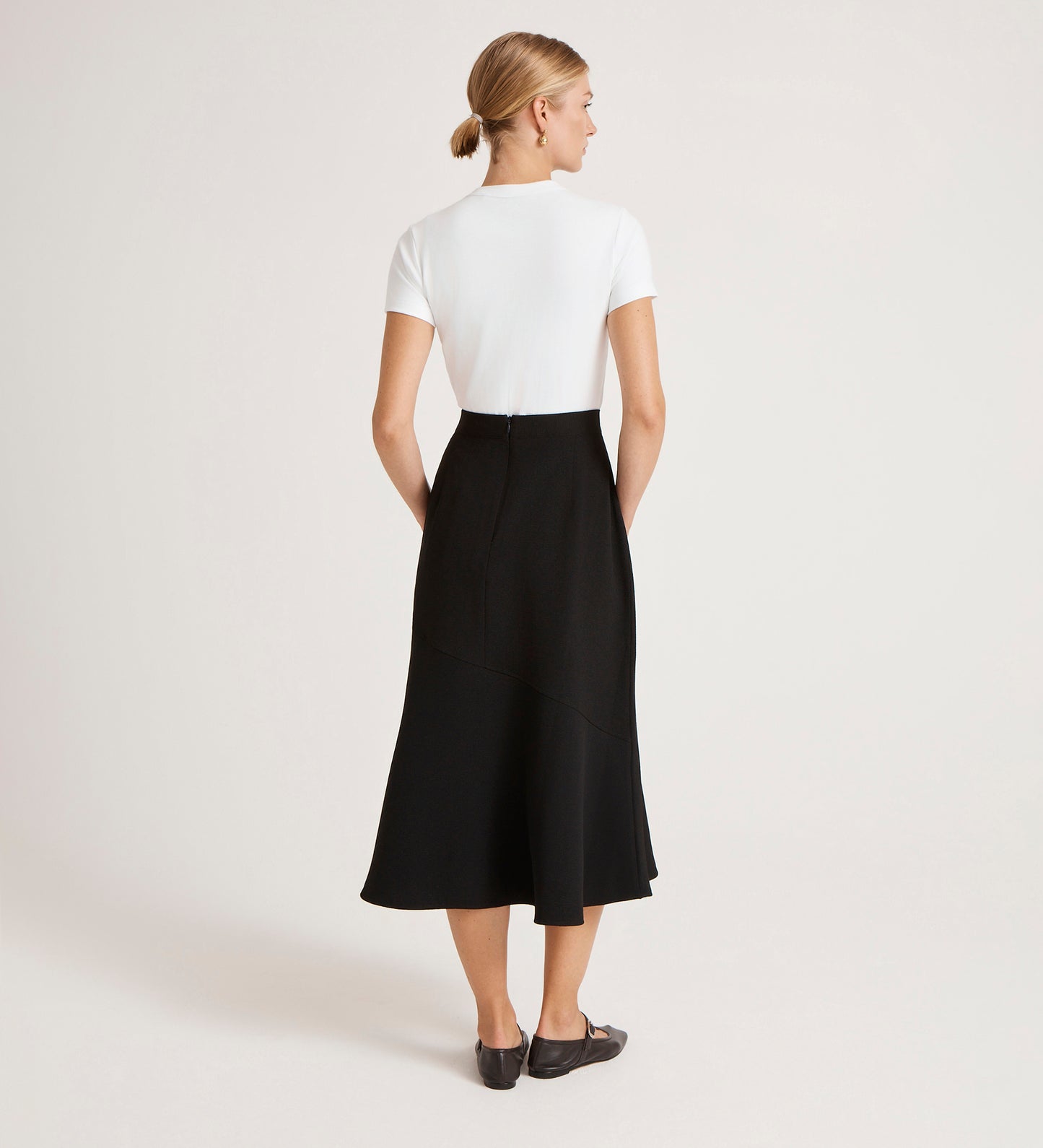 A woman with blond hair in a low bun stands with her back to the camera, wearing a white short-sleeve top tucked into the Gracie Black Rich Crepe Midi Skirt and black shoes. The skirt’s flared silhouette contrasts with the plain, light background.