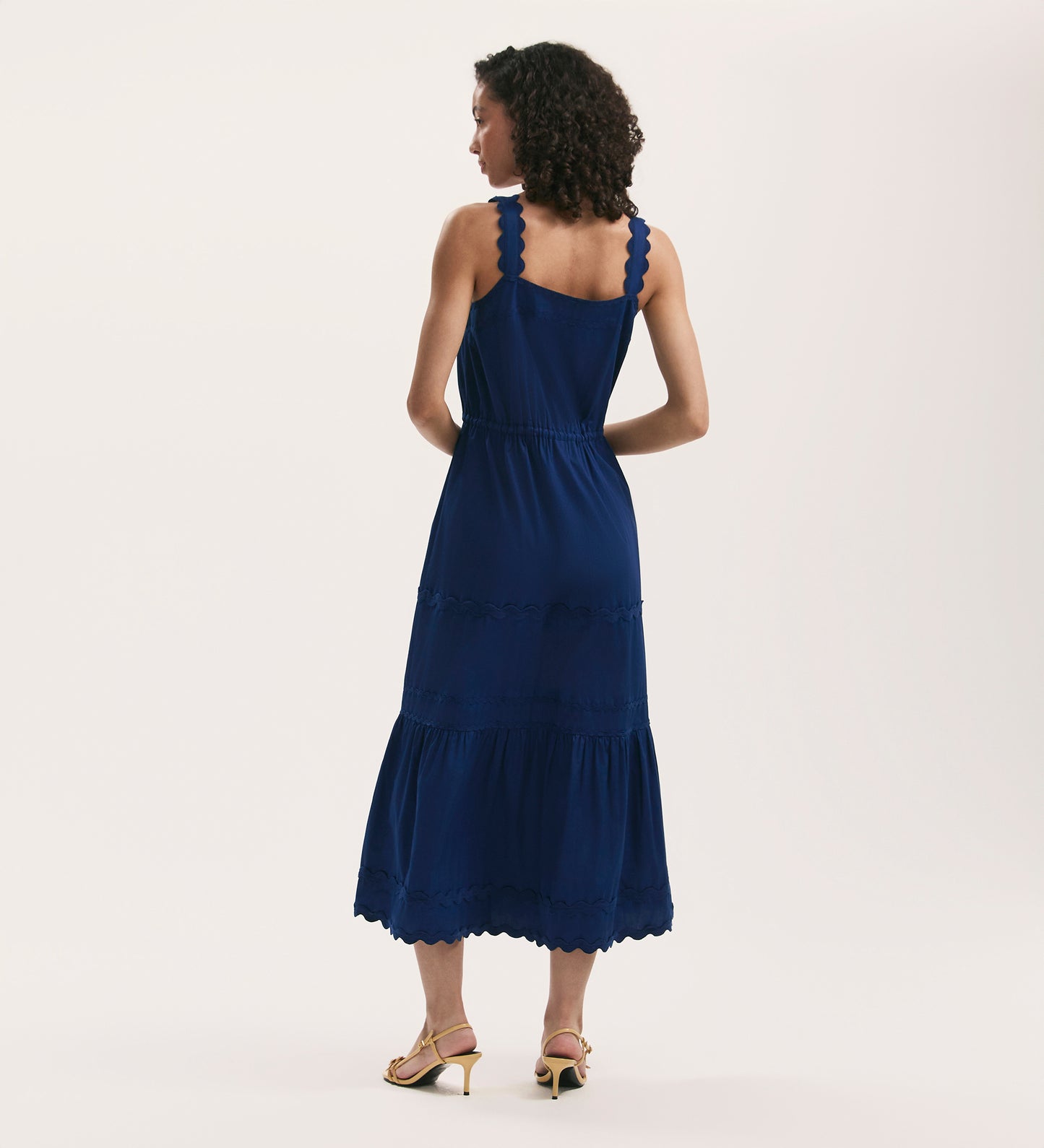 A woman with curly hair wears the Hallie Indigo Ric Rac Trim Cotton Midi Dress, standing facing away in light heeled sandals against a plain, light background.