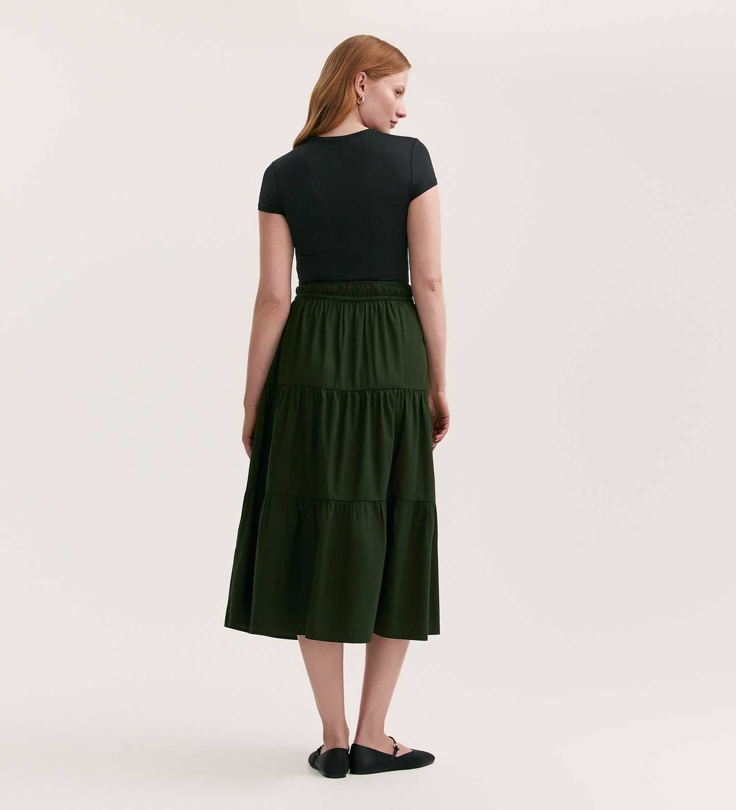 A woman stands facing away, wearing a short-sleeve black top and the Harleen Dark Khaki Cotton Midi Skirt with an elasticated waistband and tiers, paired with black flats against a plain, light-colored background.