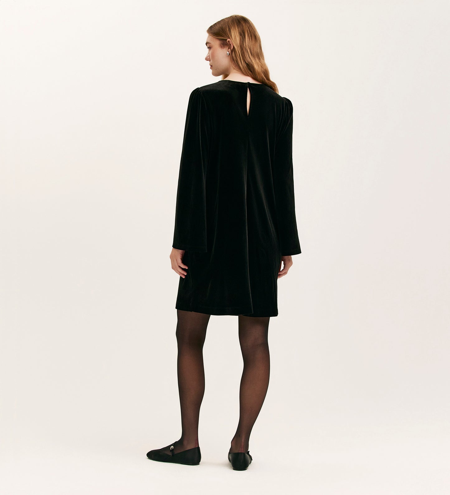 A woman with light brown hair faces away, wearing the Hattie Black Velvet Short Dress with long sleeves, paired with black sheer tights and flats, set against a plain light background.