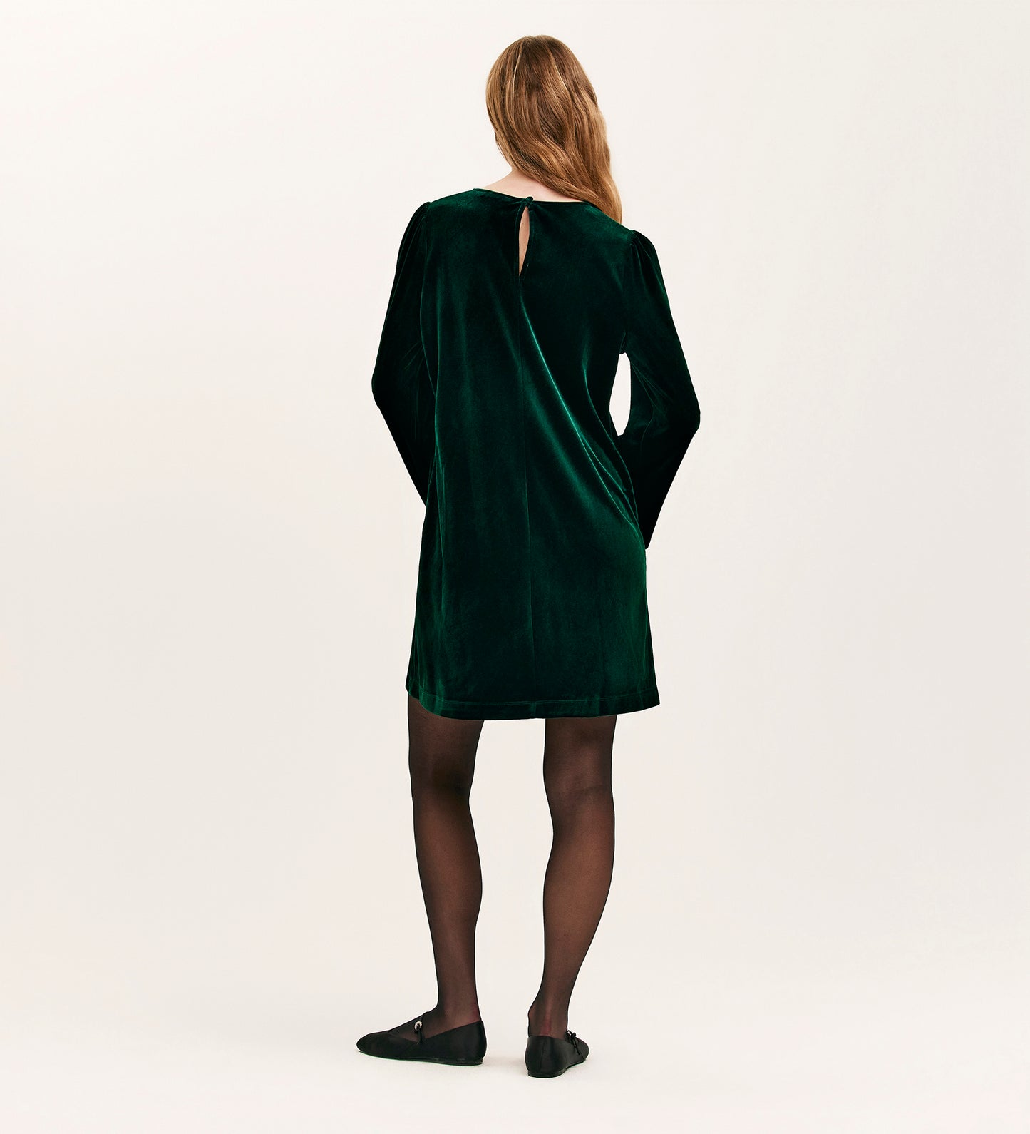 A woman with long, light brown hair faces away, wearing the Hattie Bottle Green Velvet Short Dress with long sleeves, paired with black sheer tights and flat shoes—an ideal choice for effortless party dressing.