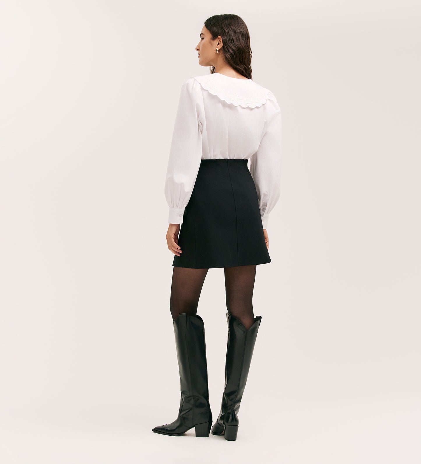 A woman stands facing away against a plain light background, wearing a white blouse with a scalloped collar, black tights, black knee-high boots, and the Helia Black Ponte Jersey Midi Skirt with an elasticated waistband.