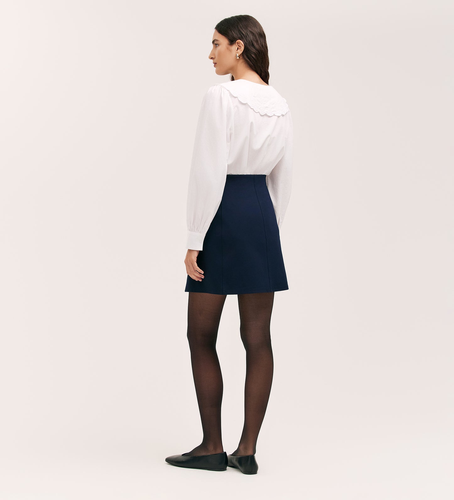 A woman stands facing away, wearing a white long-sleeve blouse with a scalloped collar, the Helia Navy Ponte Jersey Midi Skirt, black sheer tights, and black flats against a plain light background.