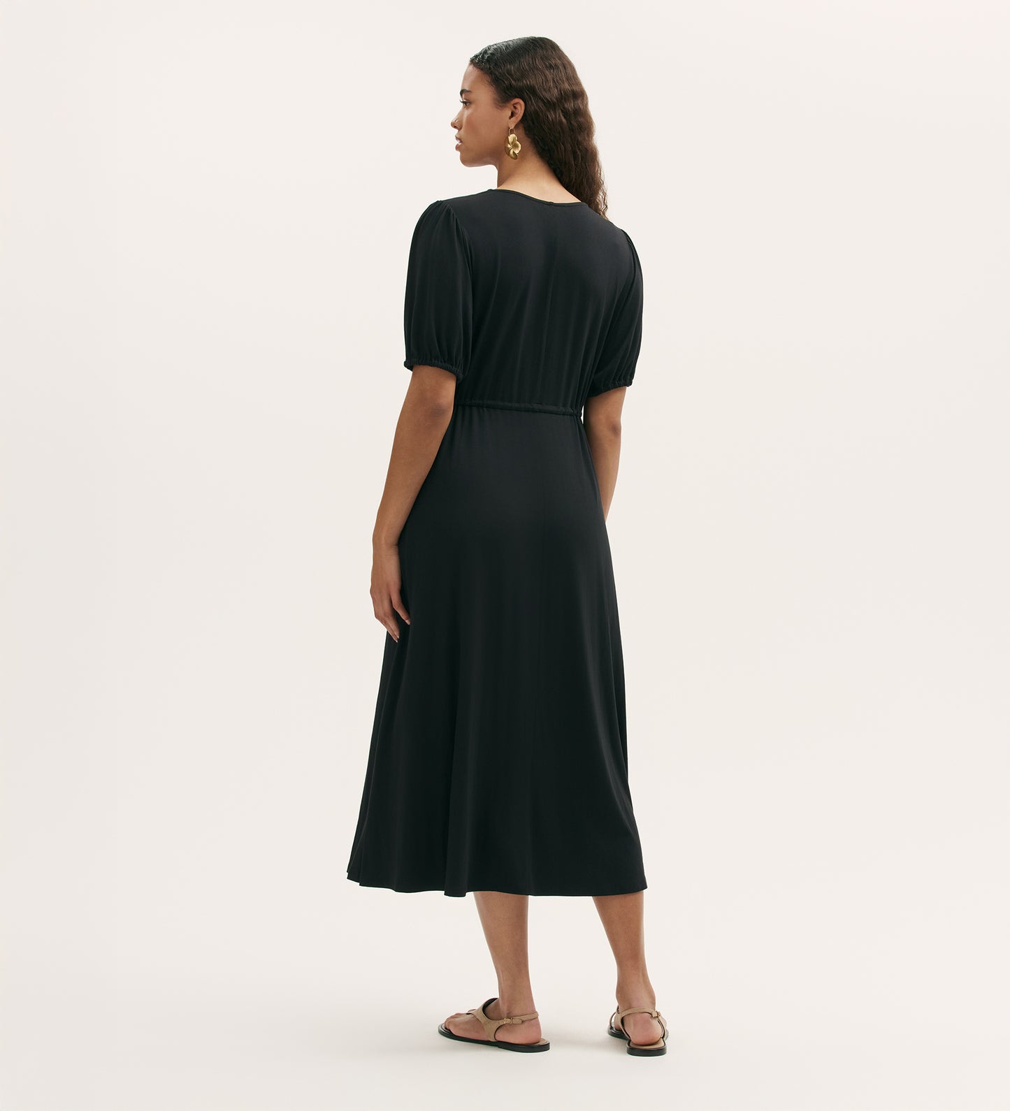 A woman with wavy hair stands facing away, wearing the Hessa Black Jersey Midi Dress featuring a v-neck and fit-and-flare silhouette. She pairs it with sandals and gold earrings against a plain light background.