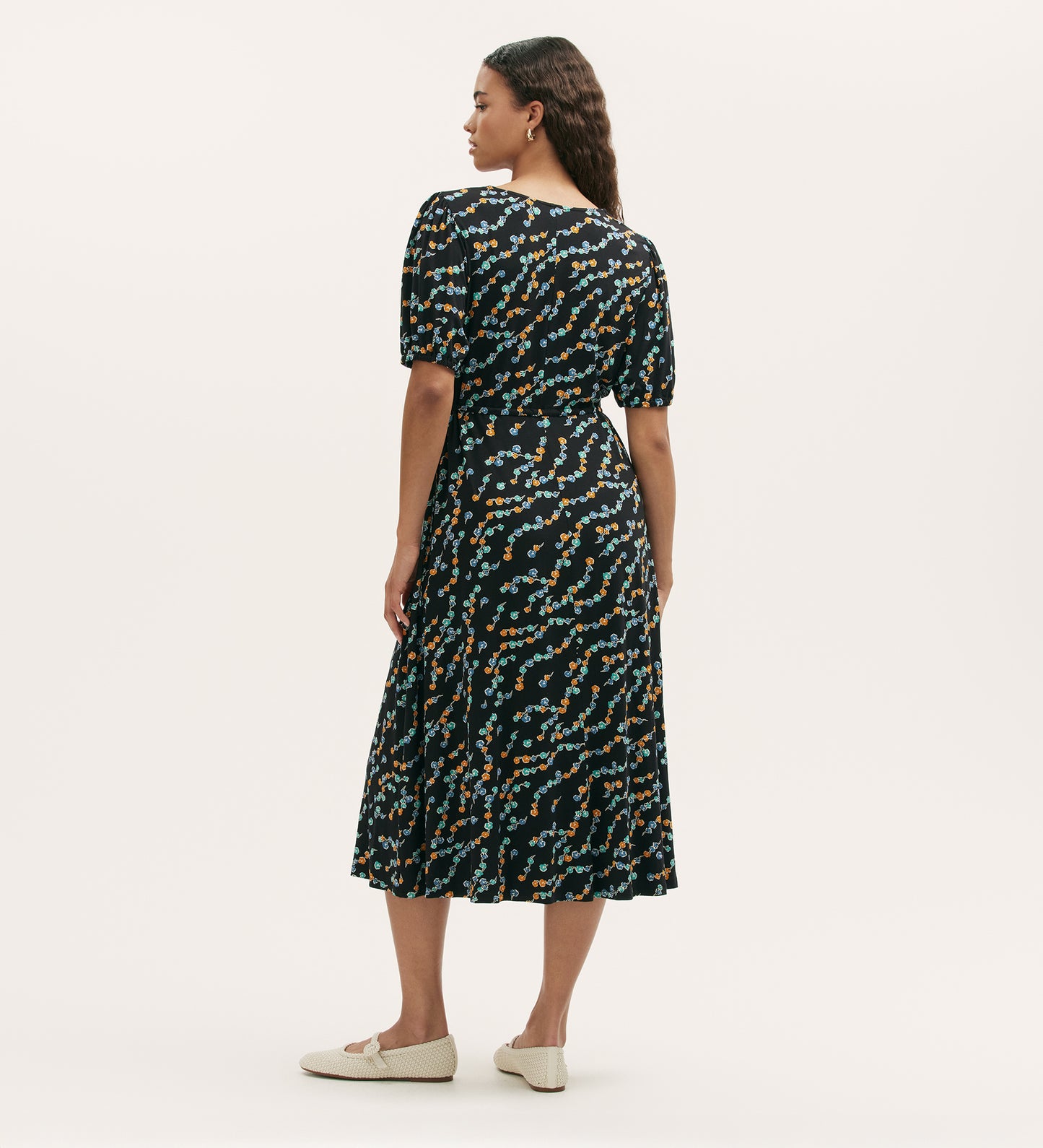 A woman faces away wearing the Hessa Black Rose Bud Jersey Midi Dress with short sleeves and a fit-and-flare silhouette, paired with beige flats, set against a plain light background.