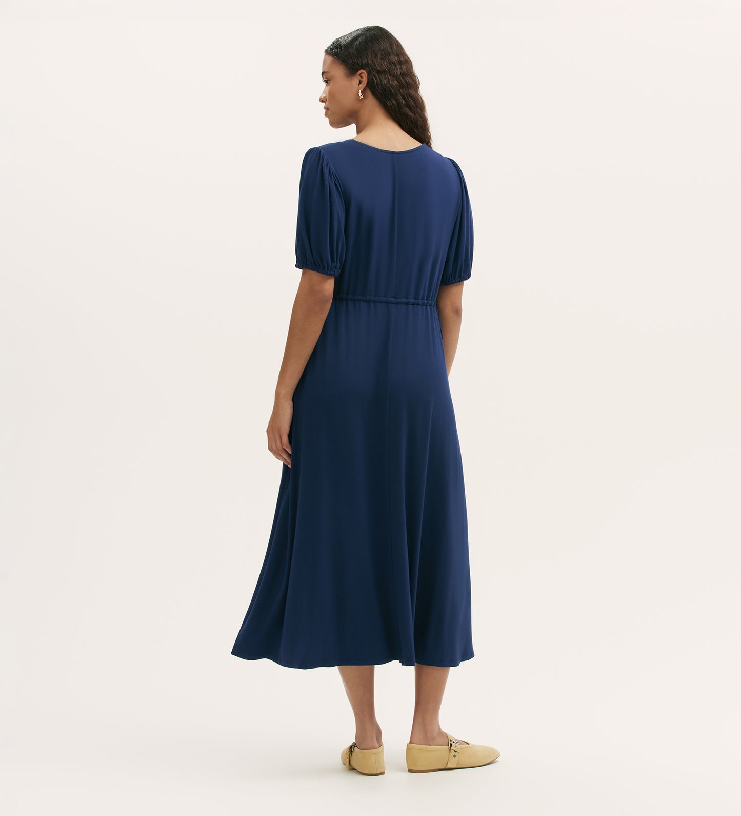Woman facing away, wearing the Hessa Navy Jersey Midi Dress with a v-neck, elastic waist, and tan flats—an effortlessly versatile staple—against a plain light background.