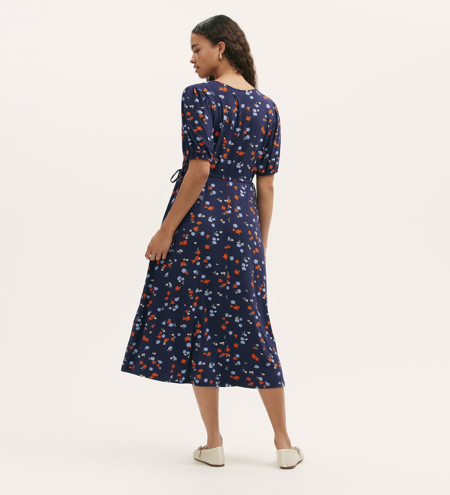 A woman with long, wavy hair wears the Hessa Navy Ditsy Jersey Midi Dress, a fit-and-flare style with short sleeves and ankle-grazing length. She pairs it with white slip-on shoes, standing against a plain background.