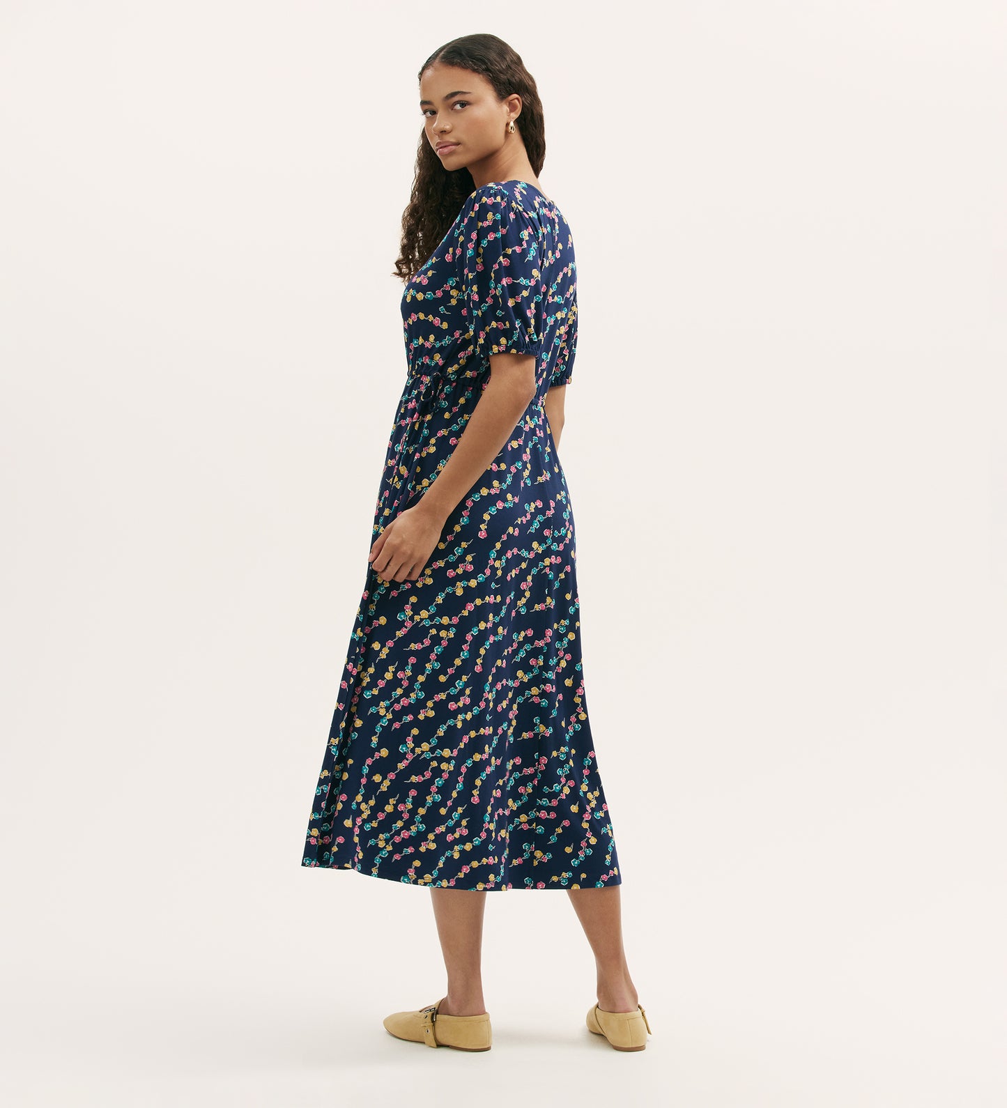 A woman in the Hessa Navy Rose Bud Jersey Midi Dress with a fit-and-flare silhouette stands sideways, looking over her shoulder. Her long, curly hair and beige shoes make this style a versatile wardrobe staple against a light background.