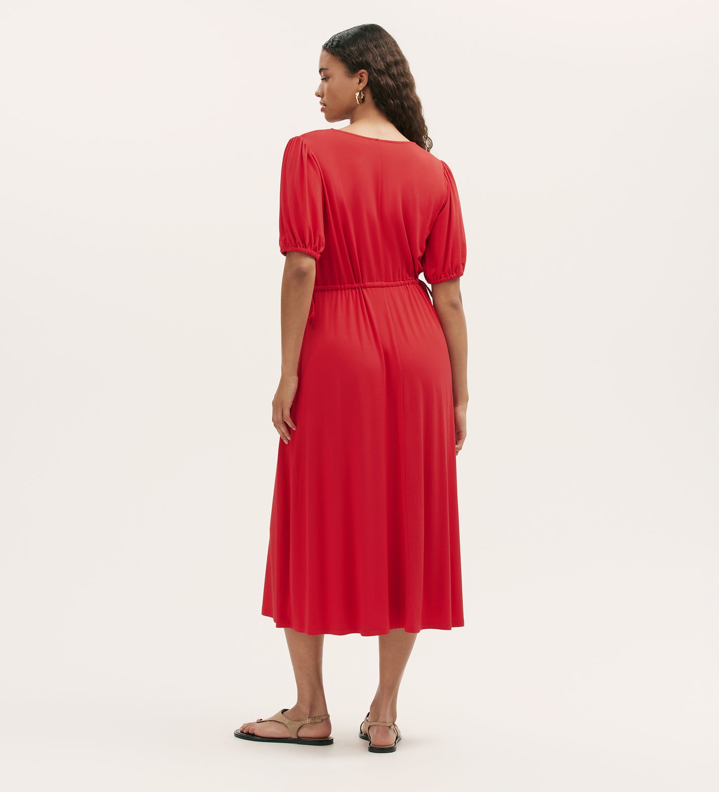 A woman faces away, wearing the Hessa Red Jersey Midi Dress with a bright red v-neck, relaxed fit, and elastic waist. She has long wavy hair and flat sandals, against a plain light background—a versatile wardrobe staple.