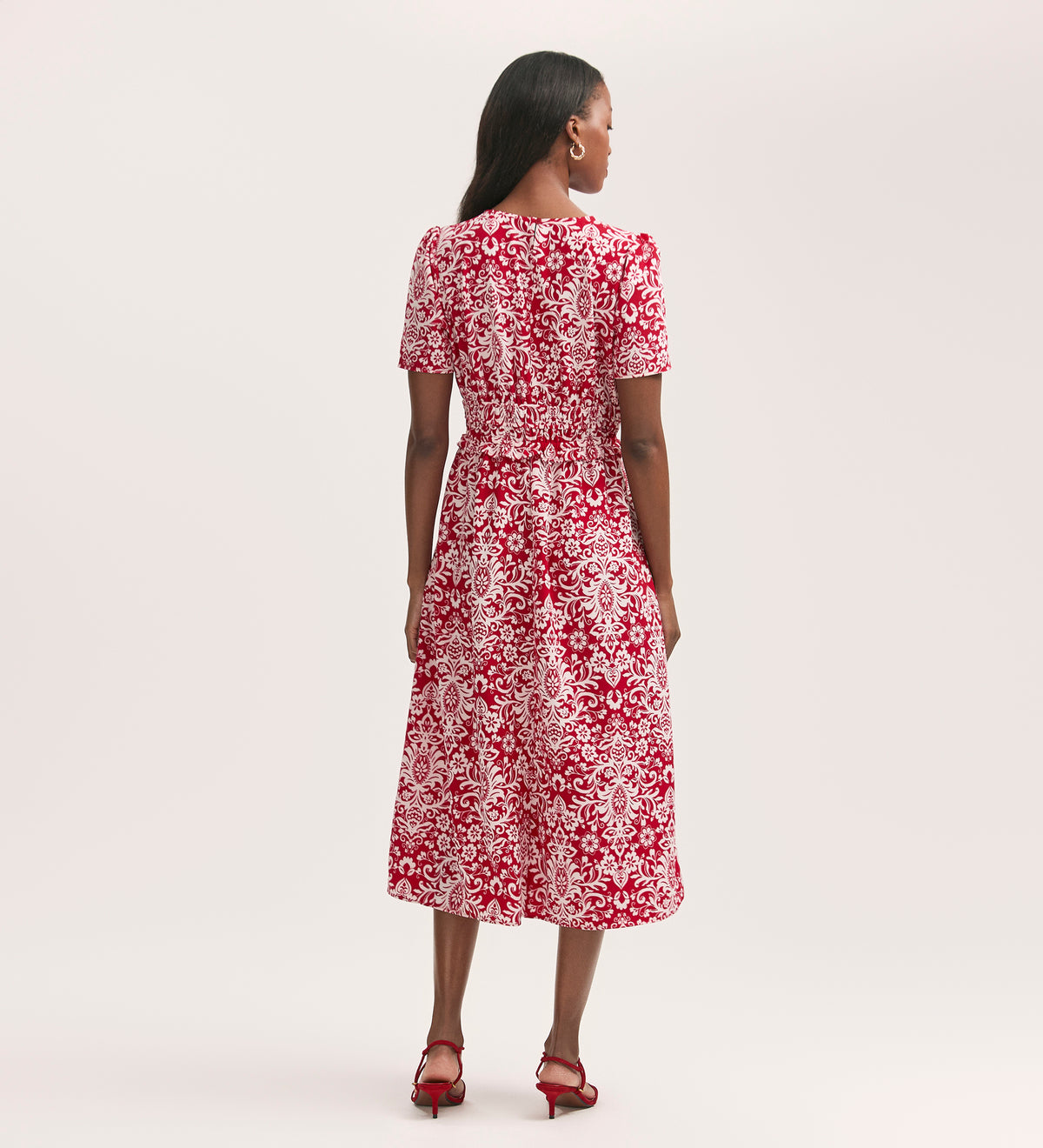 A woman stands facing away, wearing the Irini Red Paisley Crepe Midi Dress with short sleeves and an elasticated waist, paired with red heeled sandals against a plain light background.