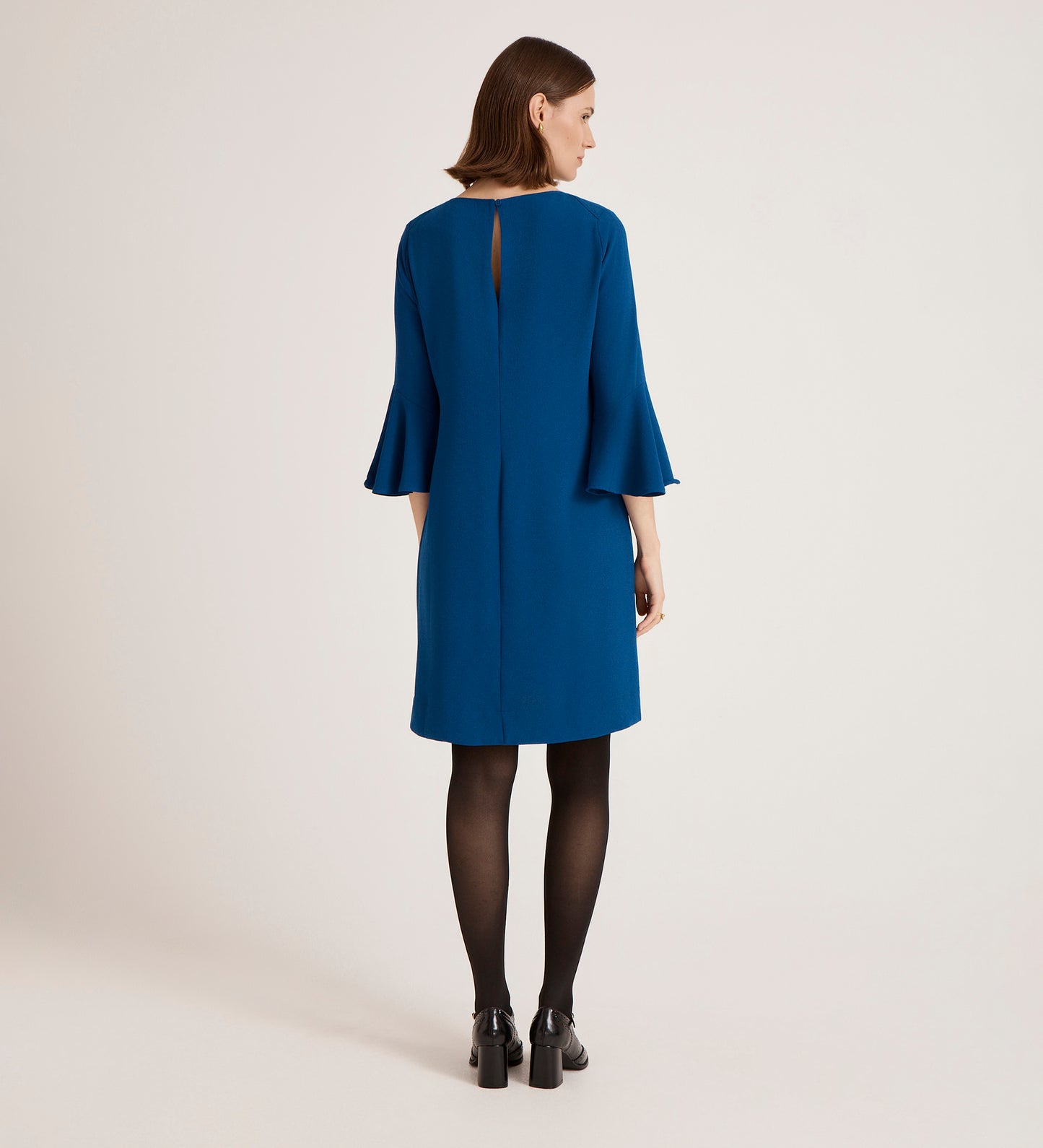 A woman with straight brown hair stands with her back to the camera, wearing a blue Izzy Cobalt Crepe Short Dress, black tights, and black shoes against a plain background.