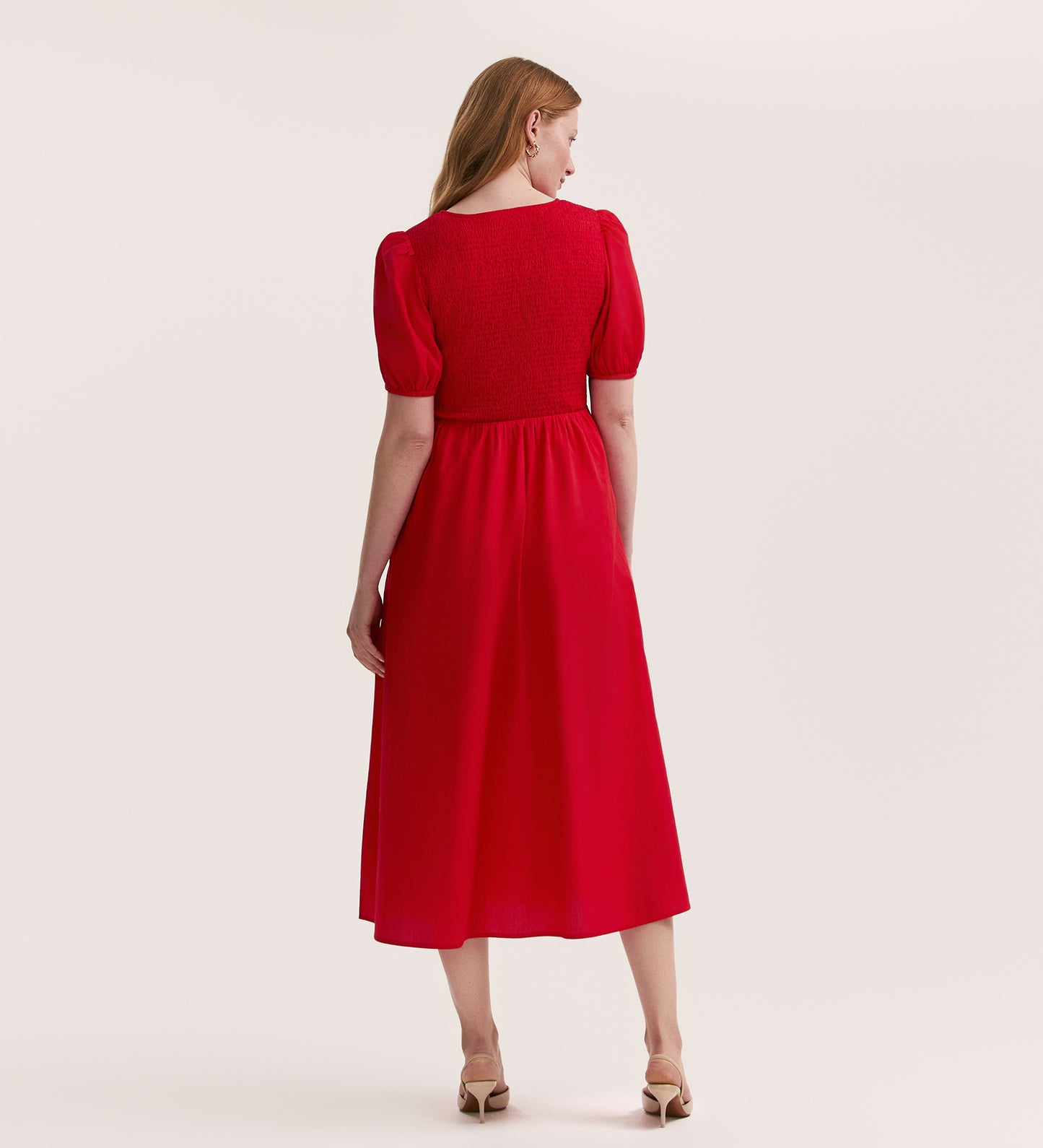 A woman with long, light brown hair stands with her back to the camera, wearing the Jamilla Red Cotton Midi Dress with a v-neckline and shirred bodice, paired with beige heels against a plain light background.