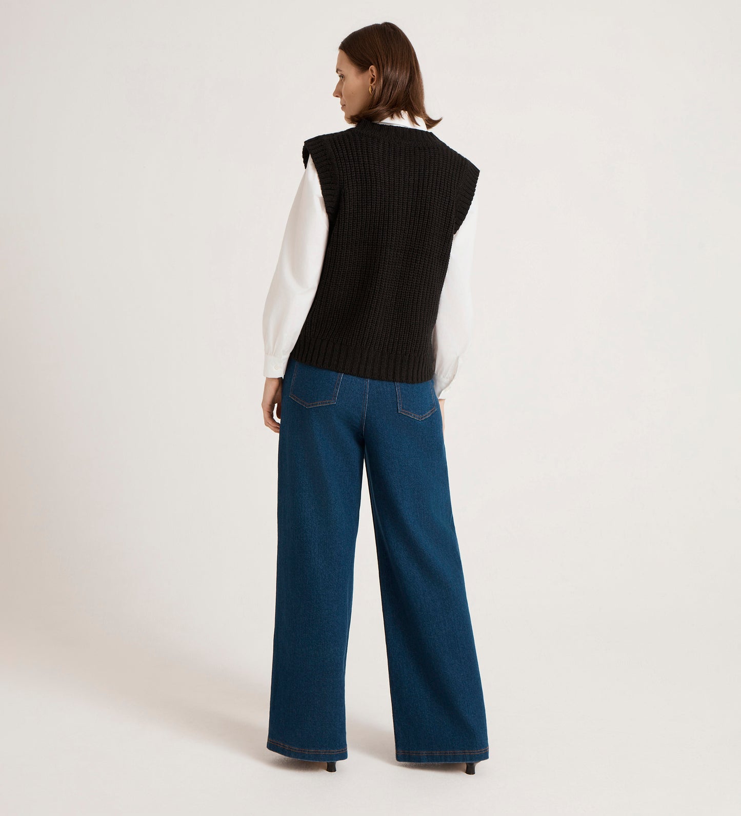 A woman faces away, wearing a white long-sleeve shirt with the Janette Black Knitted Vest layered on top and wide-leg blue jeans. Her chin-length brown hair stands out against the plain background for an effortlessly chic look.