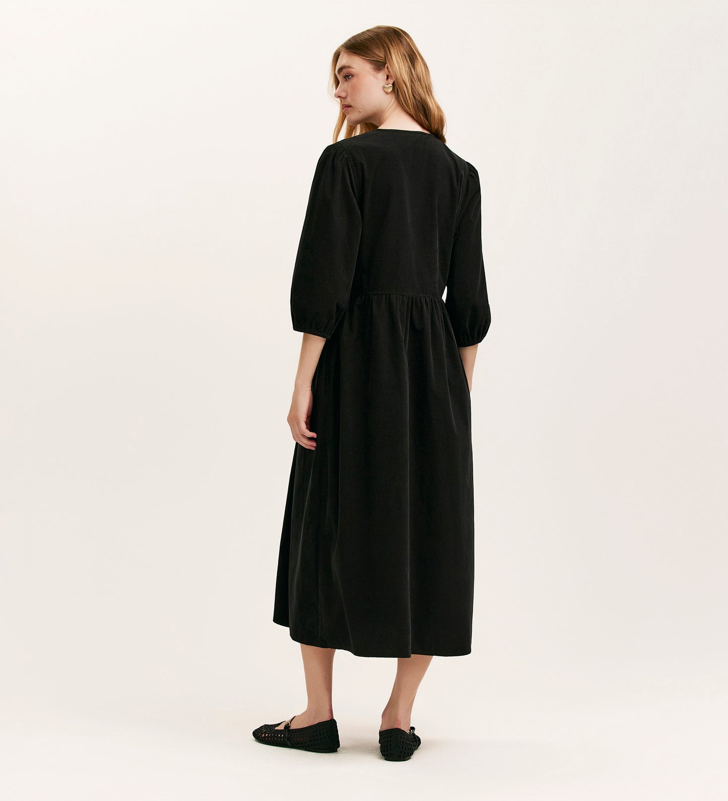 A woman with long, light brown hair faces away, wearing the Jaz Black Corduroy Midi Dress with three-quarter sleeves and black flats, posed against a plain light background.