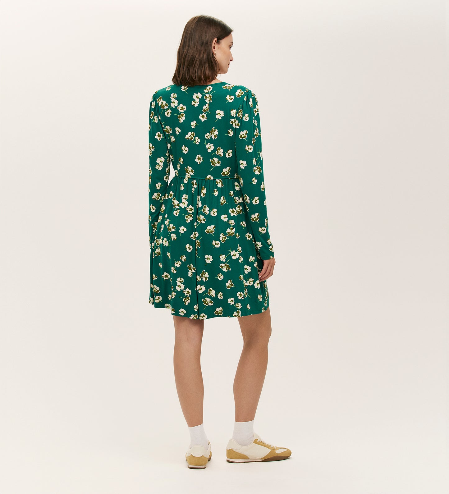 A woman faces away, wearing the Joanne Green Poppies Jersey Short Dress, paired with white socks and yellow shoes, set against a plain light background.