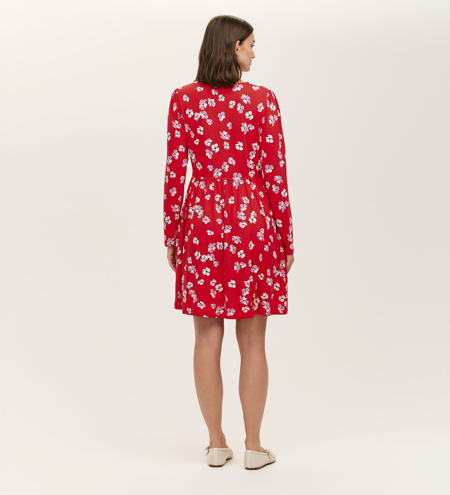 A woman faces away, wearing the Joanne Red Poppies Jersey Short Dress with white floral print and beige flats, ideal for everyday comfort. The background is plain and light-colored.