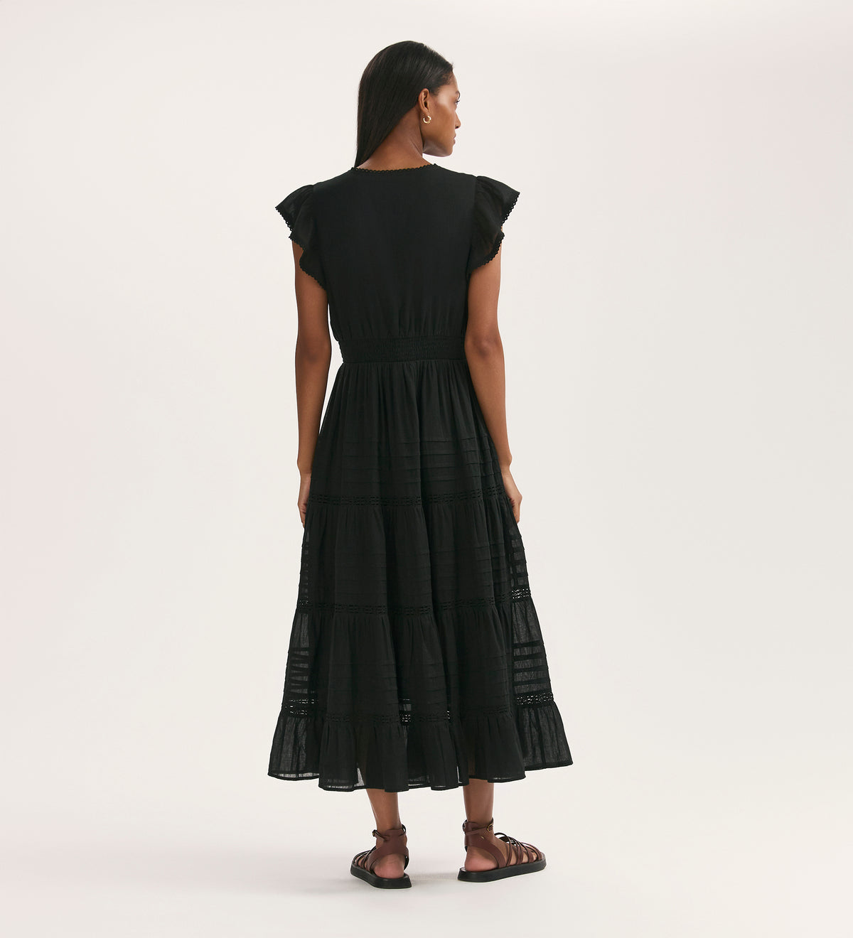 A woman faces away, wearing the Jude Black Cotton Midi Tiered Dress with ruffled sleeves and sandals. The cotton fabric and fitted waist enhance its flowing, ankle-length skirt. The background is plain and light-colored.