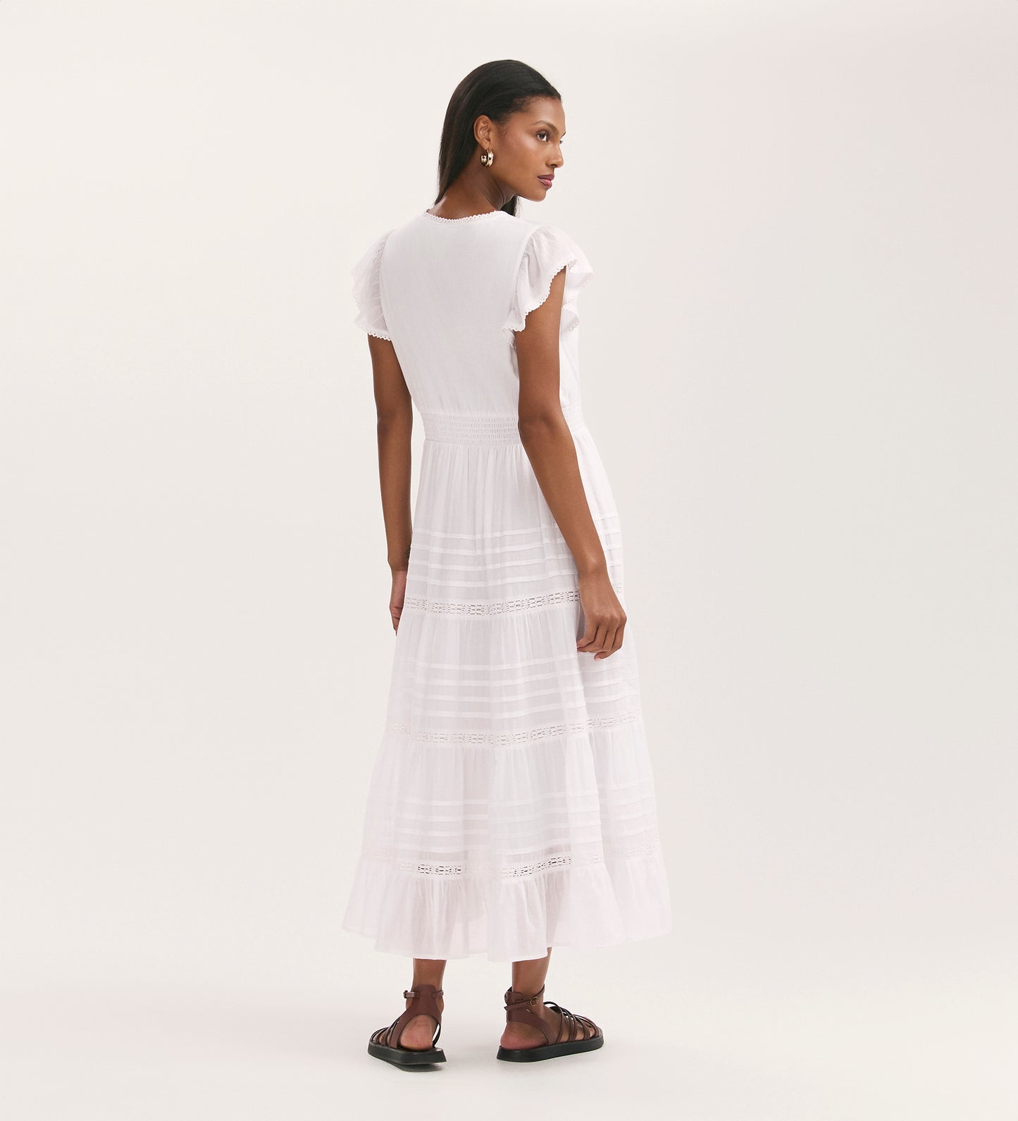 A woman faces away, wearing the Jude White Cotton Midi Tiered Dress with scallop lace trim and flat brown sandals, set against a plain light background.